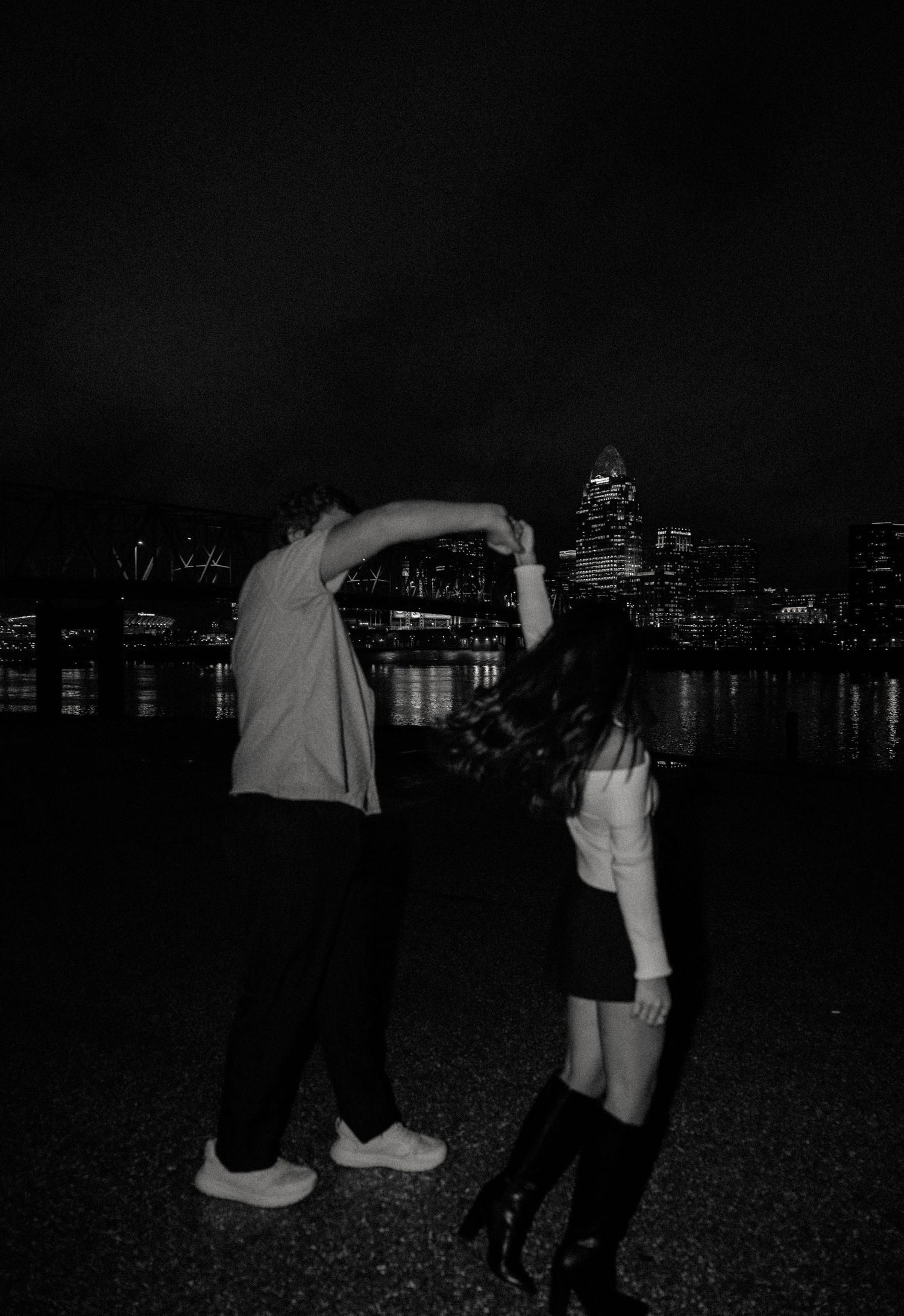 Couple dancing together at night by a city waterfront skyline in a romantic black and white photo.