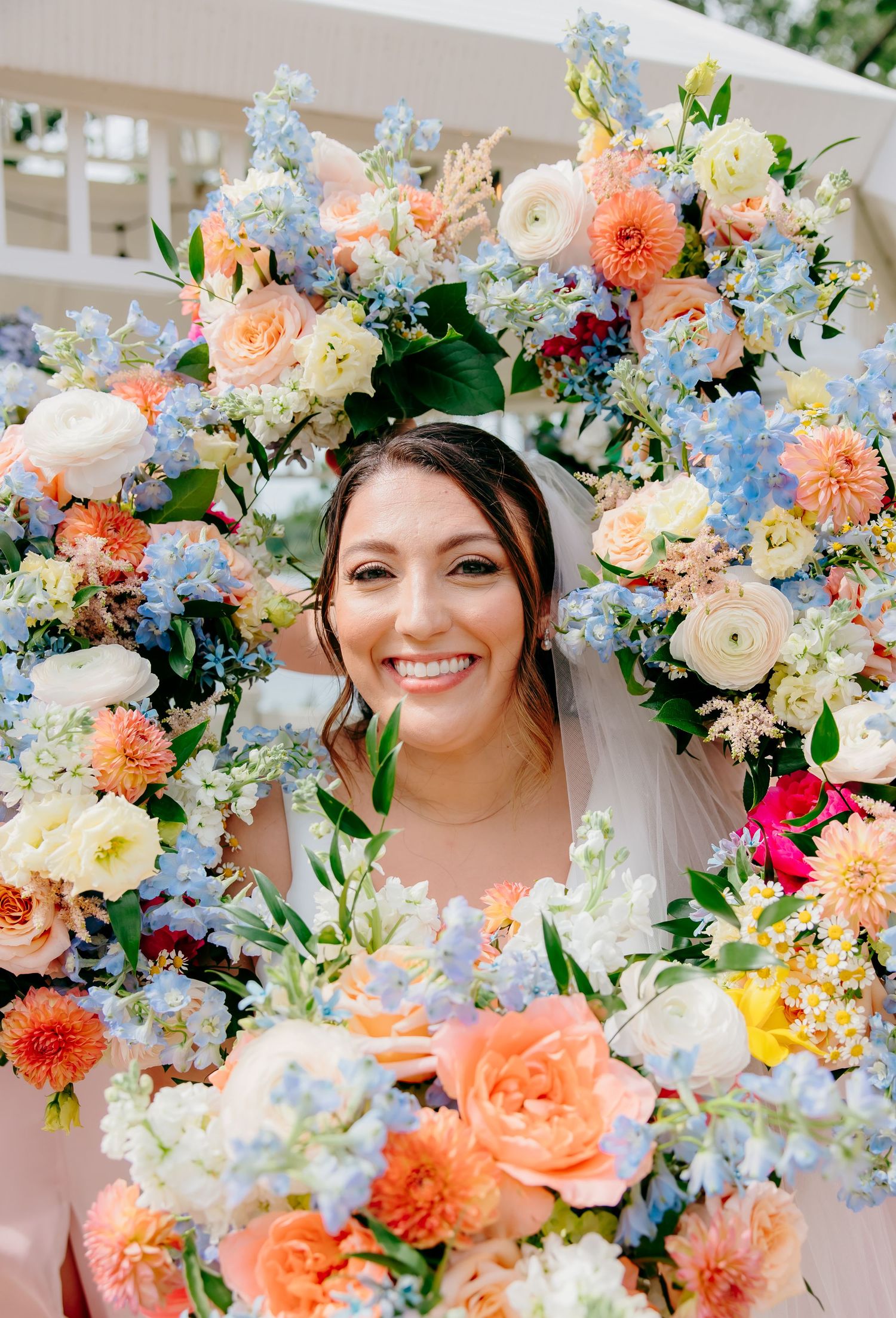 Smiling bride framed by a colorful floral wreath of blue, peach, and white blooms on her wedding day.