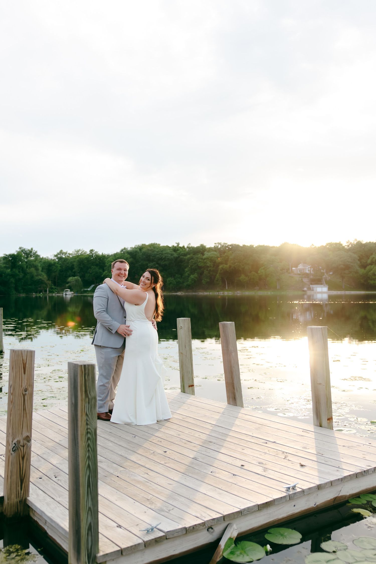 Bride and groom embrace on a wooden dock at sunset, with a serene lake and lush trees reflecting golden light behind them.