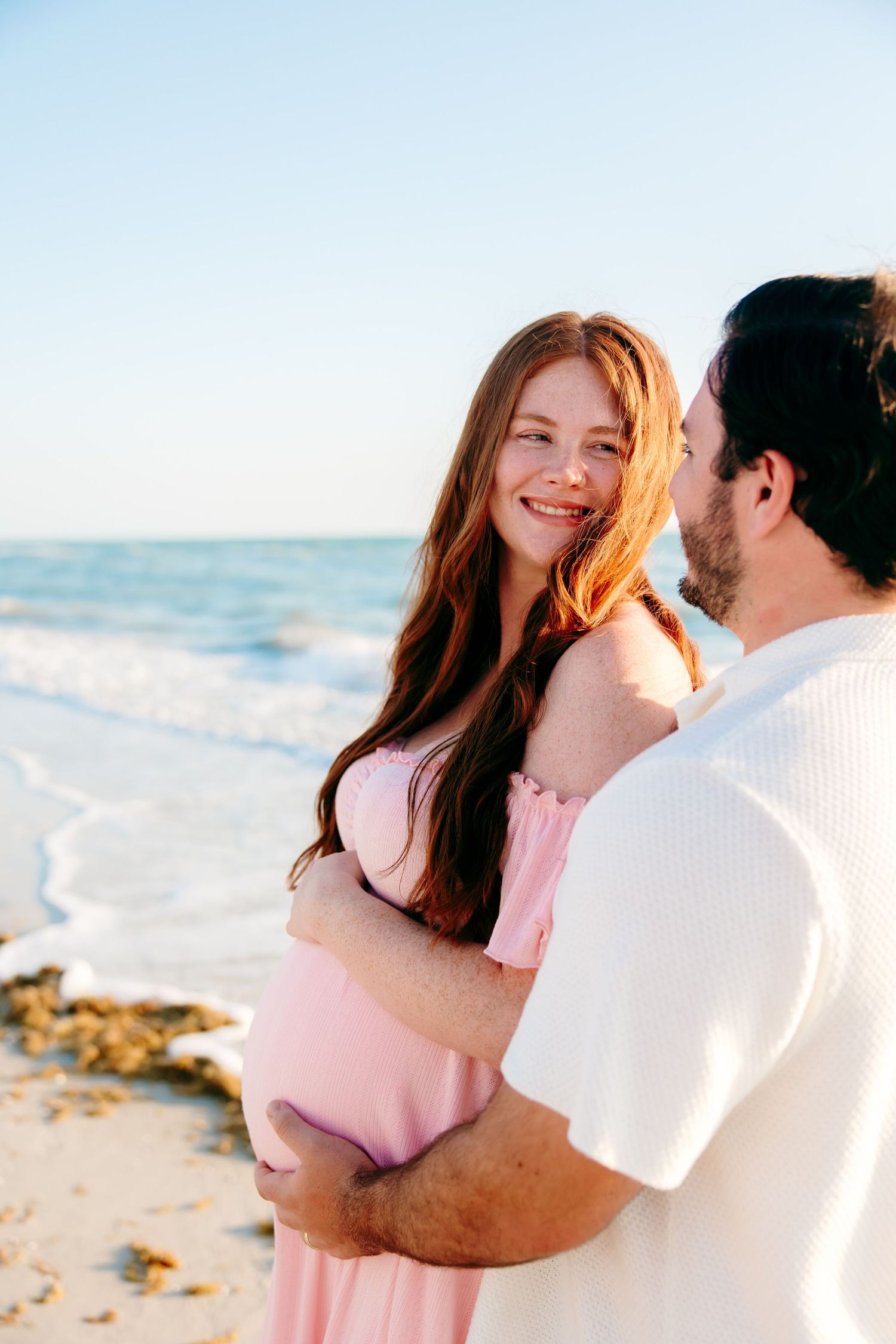 Pregnant woman in pink dress smiles at partner while embracing on sunny beach with ocean waves.