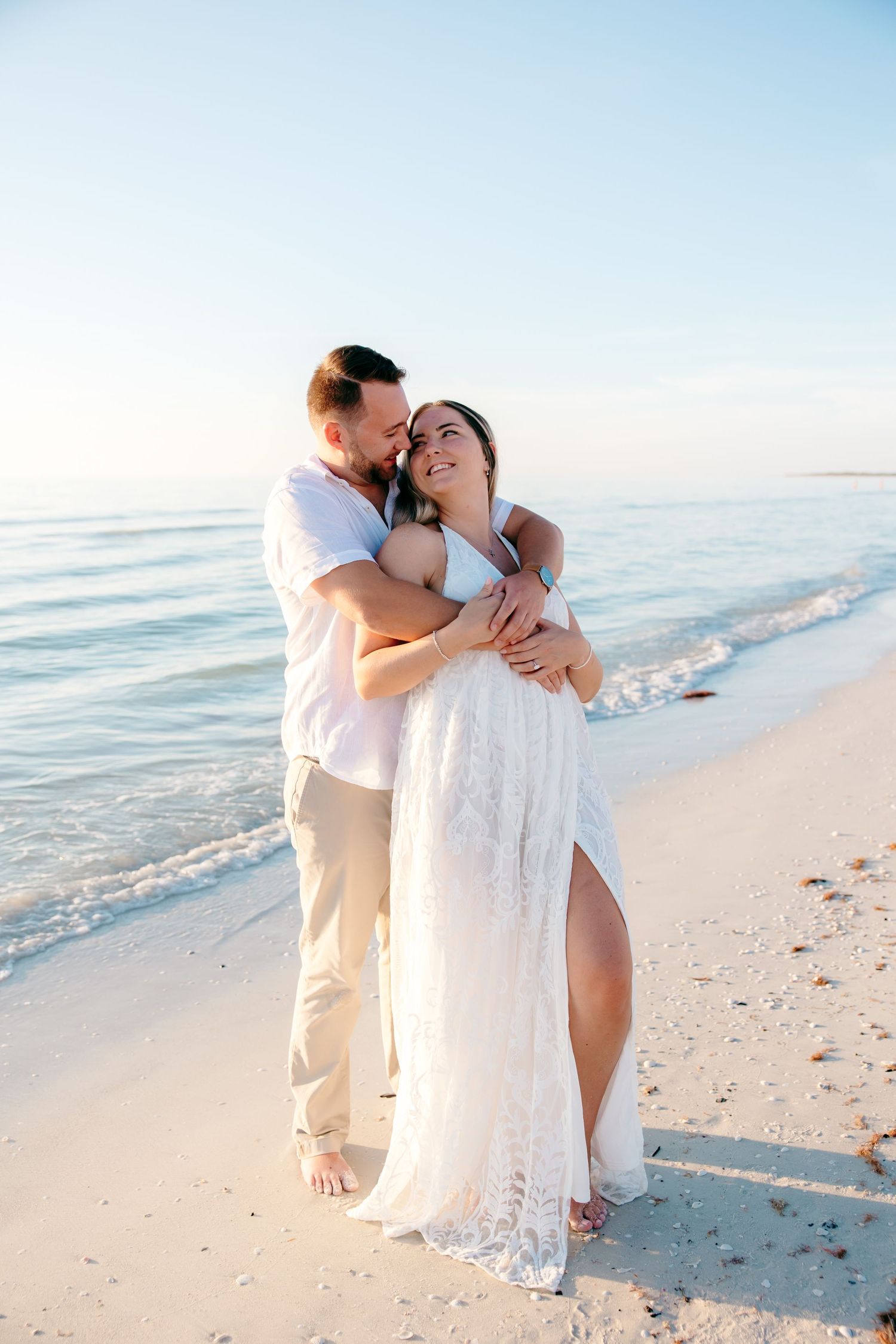 A romantic couple embraces on a pristine white sandy beach at sunset with gentle waves in the background.