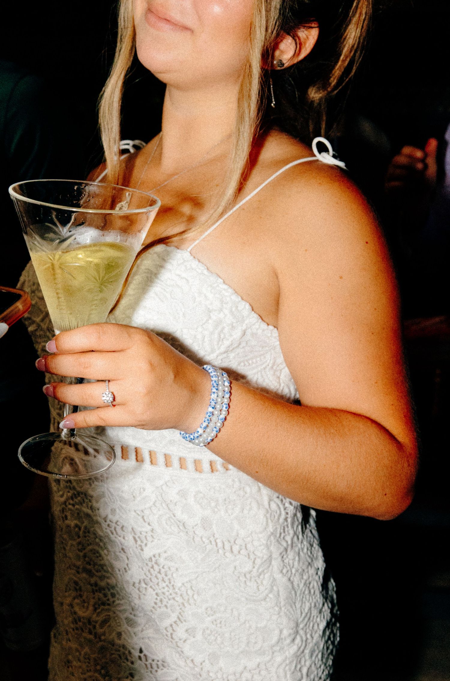 Smiling bride in white lace dress holding champagne martini glass with diamond ring and bracelet.