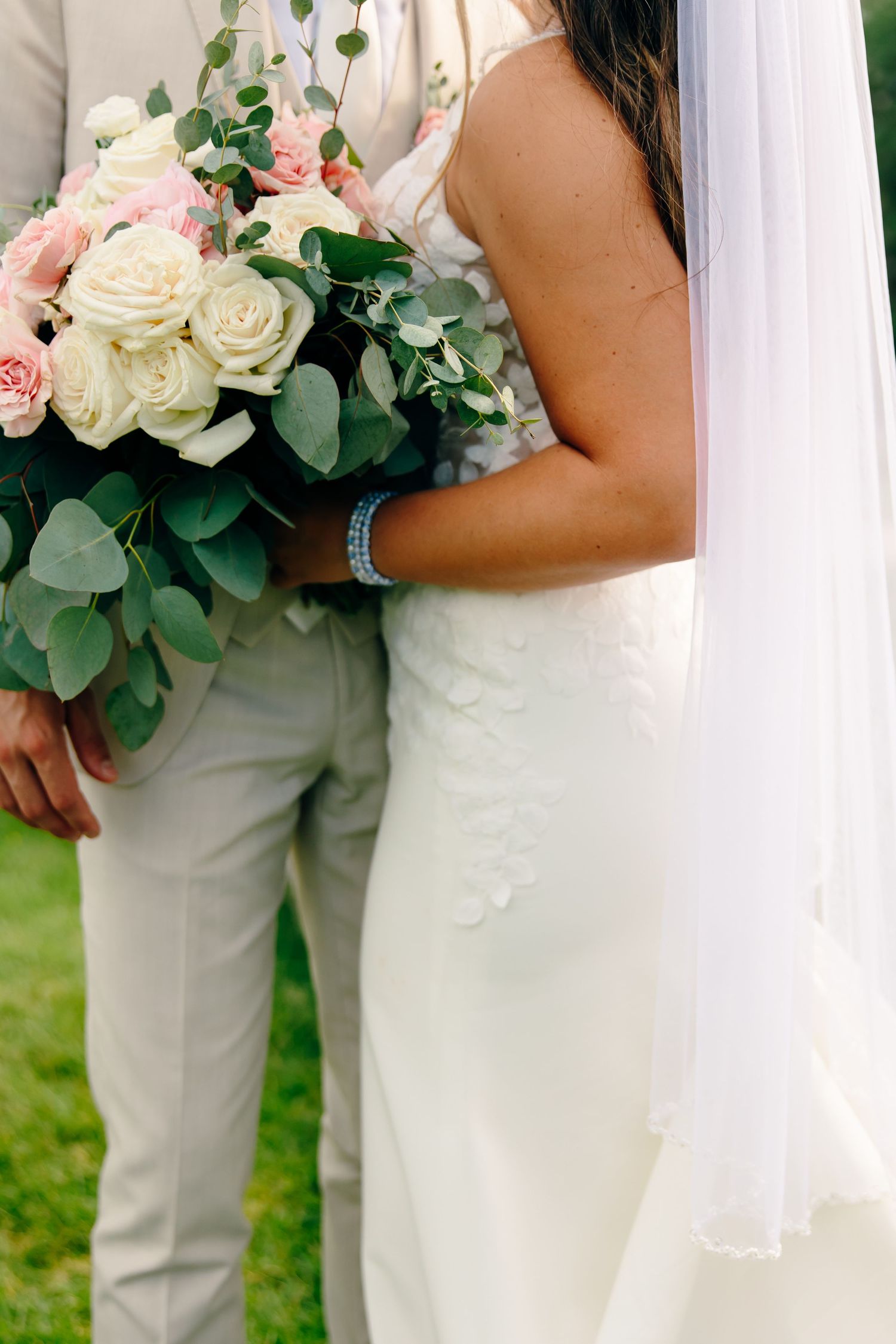 Bride in white lace gown holding pink and white rose bouquet with eucalyptus, standing beside groom in tan suit.