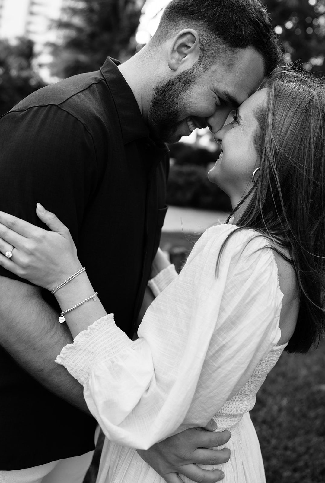 Black and white photo of a smiling couple embracing closely, woman in white dress with ring visible.