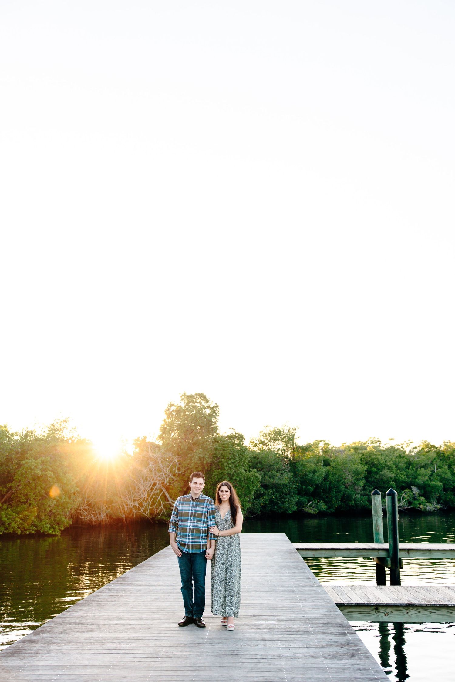 Couple holding hands on a wooden dock at golden hour sunset with lush green trees and calm water reflections.