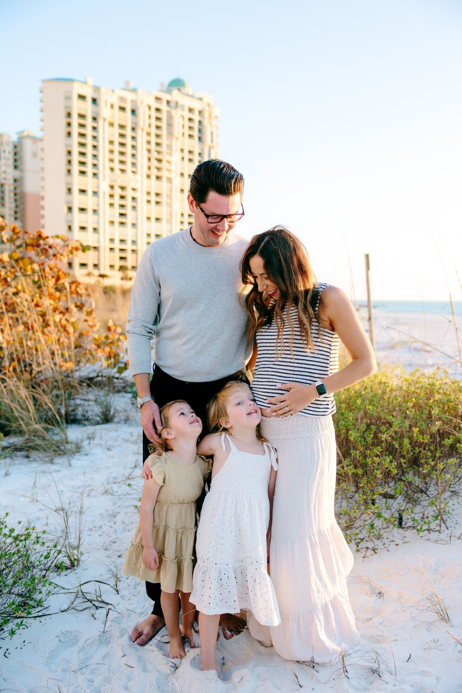 Family of four poses on sandy beach with coastal building backdrop, parents smiling at two young daughters in golden light.