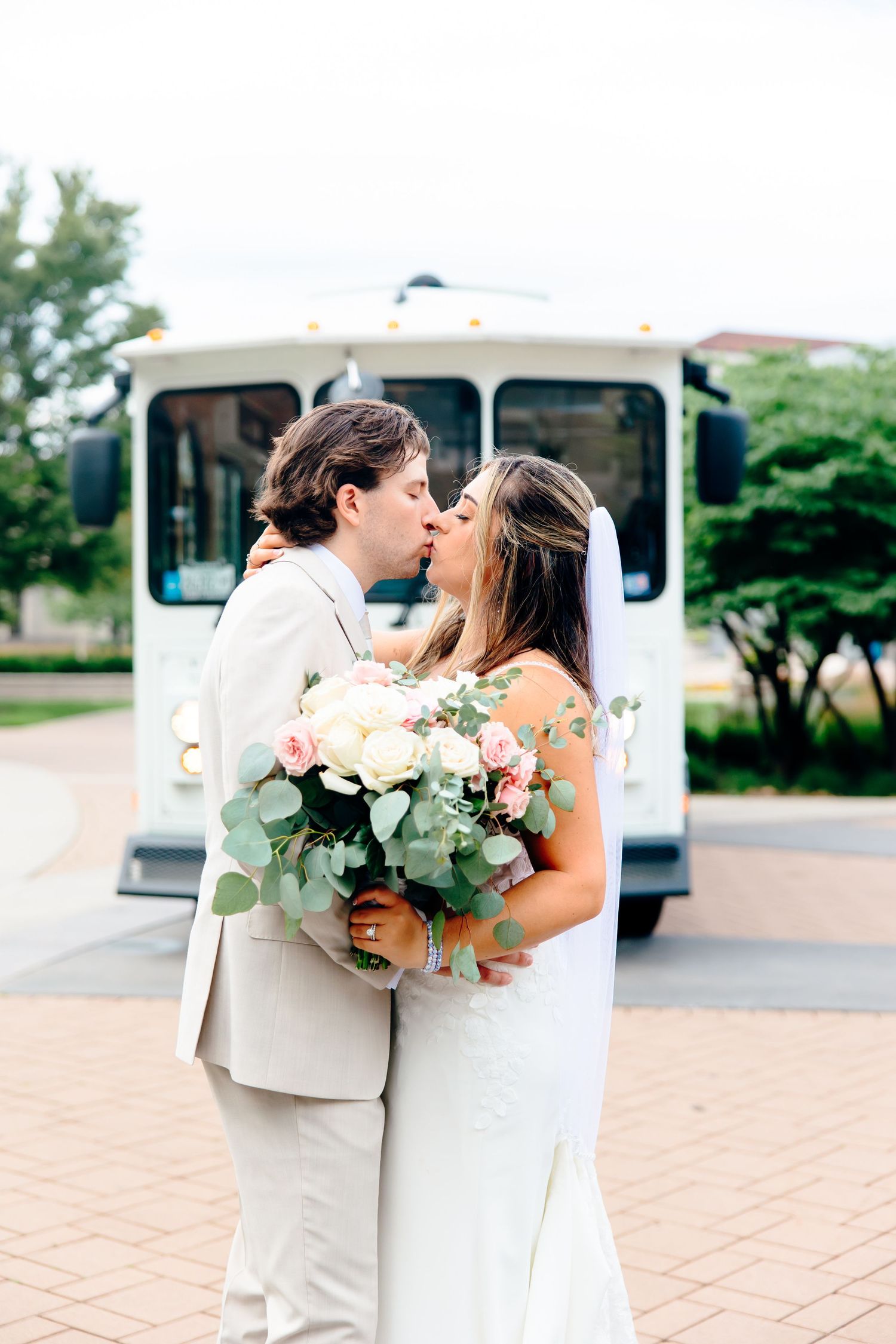 Bride and groom share a kiss holding a white and pink bouquet with a white trolley bus in the background.