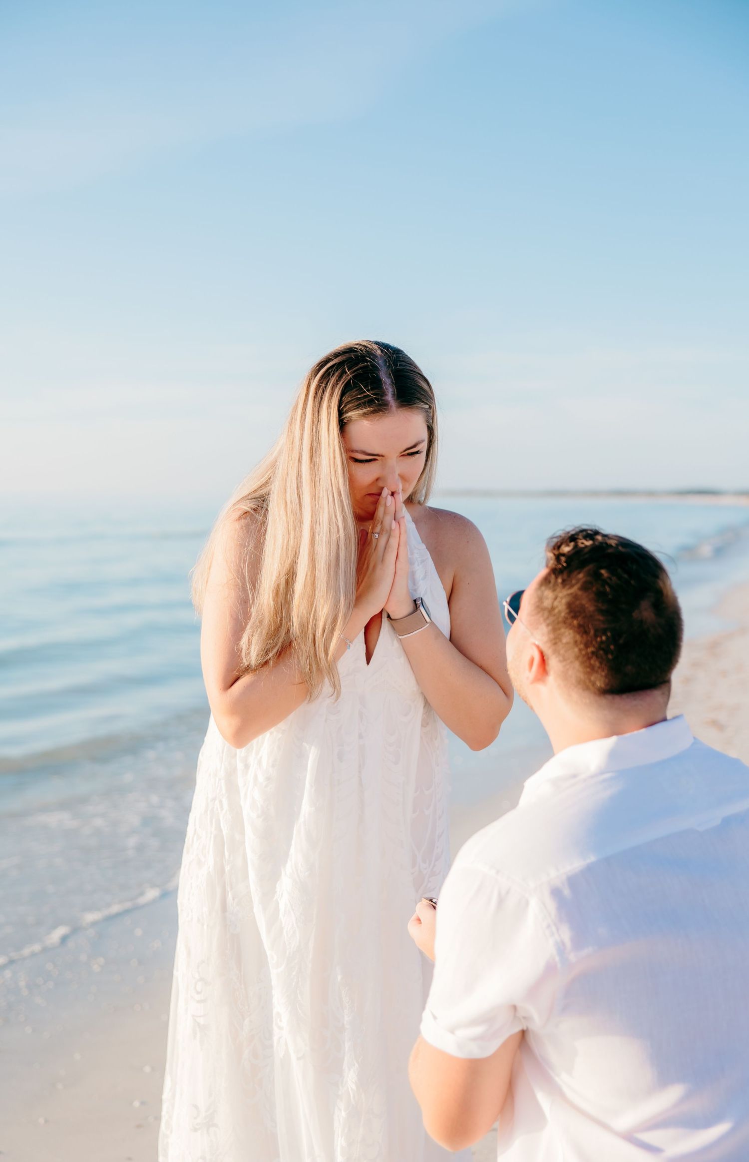 A romantic marriage proposal scene unfolds on a serene beach with soft blue ocean waters in the background.