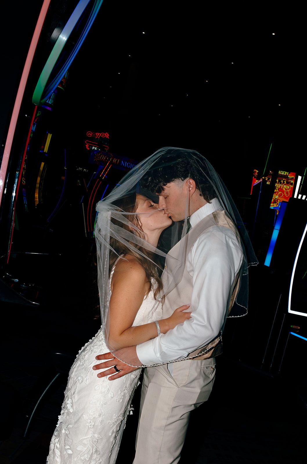 Bride and groom sharing a romantic first kiss under a veil in a colorful neon-lit venue on their wedding day.