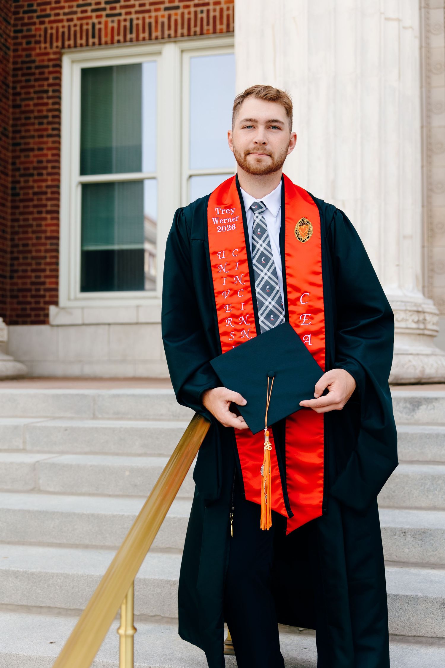 Young man in black graduation gown and red honor stole holds cap on university steps, celebrating academic achievement.