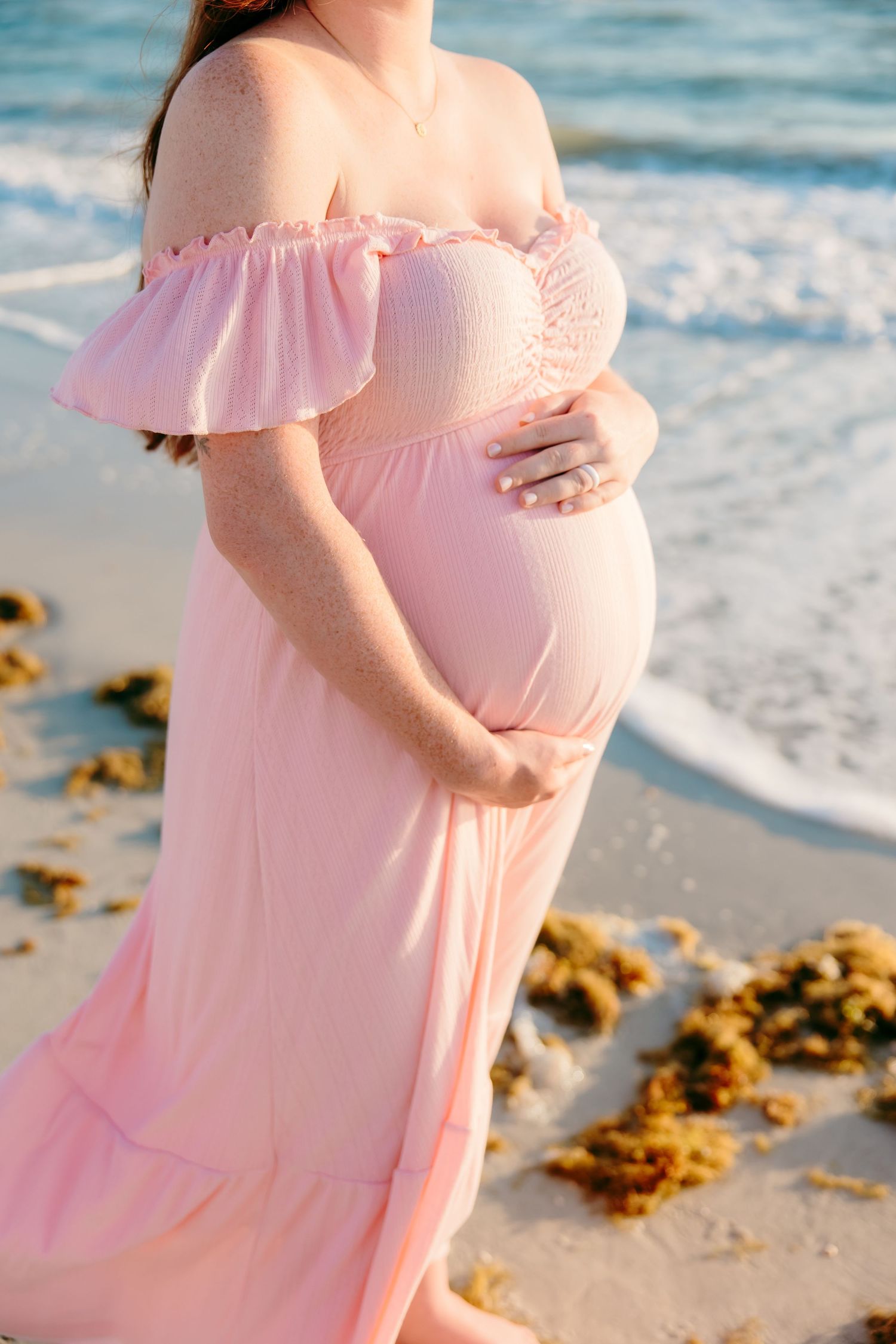 Pregnant woman in pink off-shoulder dress cradling baby bump on sandy beach with ocean waves.