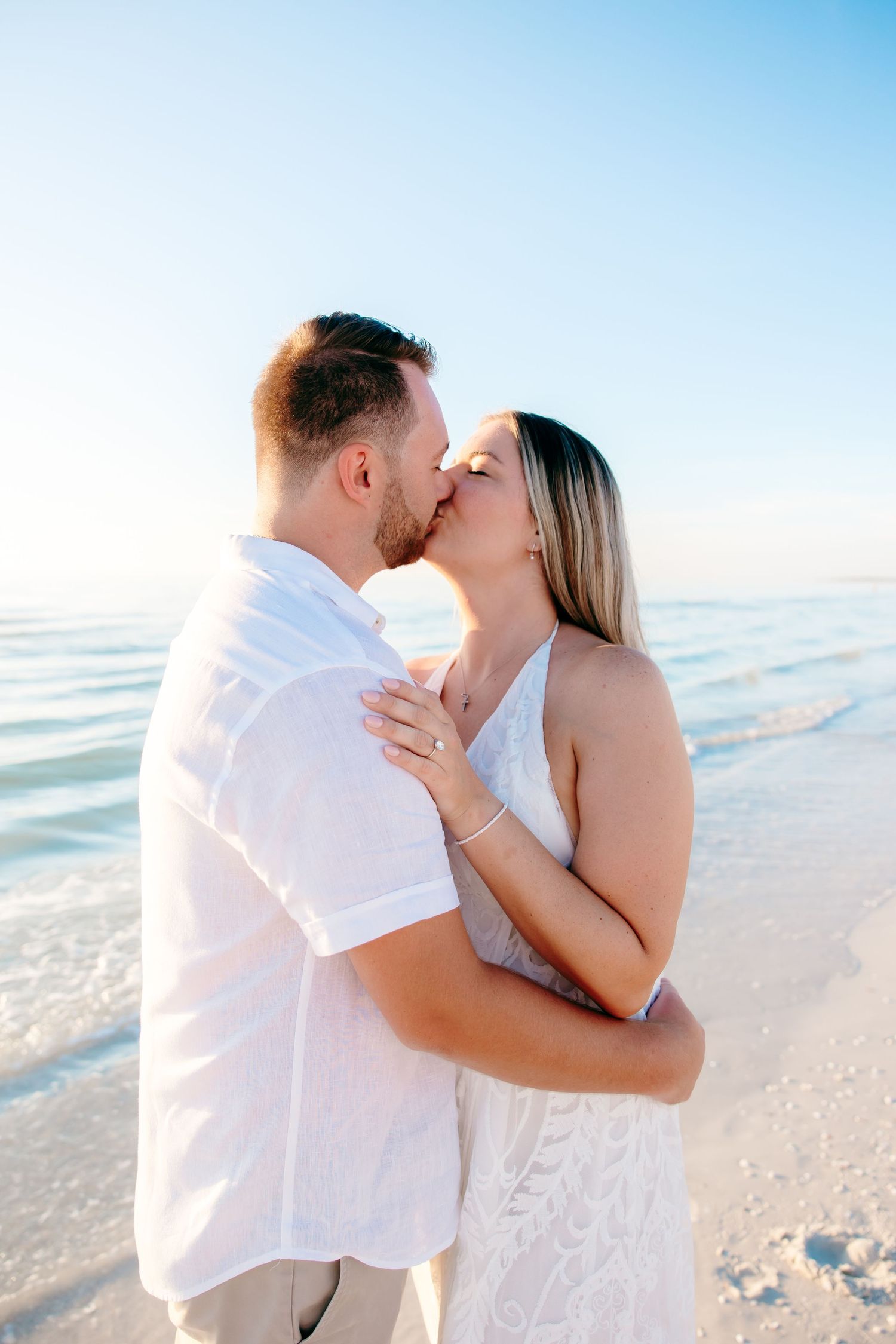 A romantic couple shares tender kisses on a sunny beach with white sand and blue ocean waves in the background.