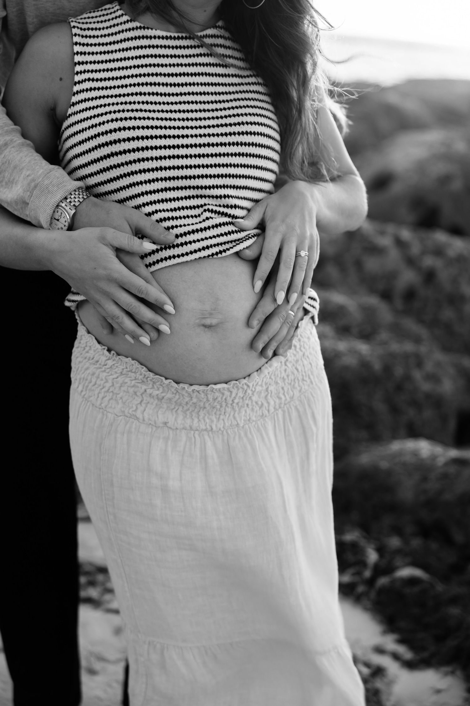 Black and white maternity photo of couple cradling pregnant belly outdoors near rocky coastline.