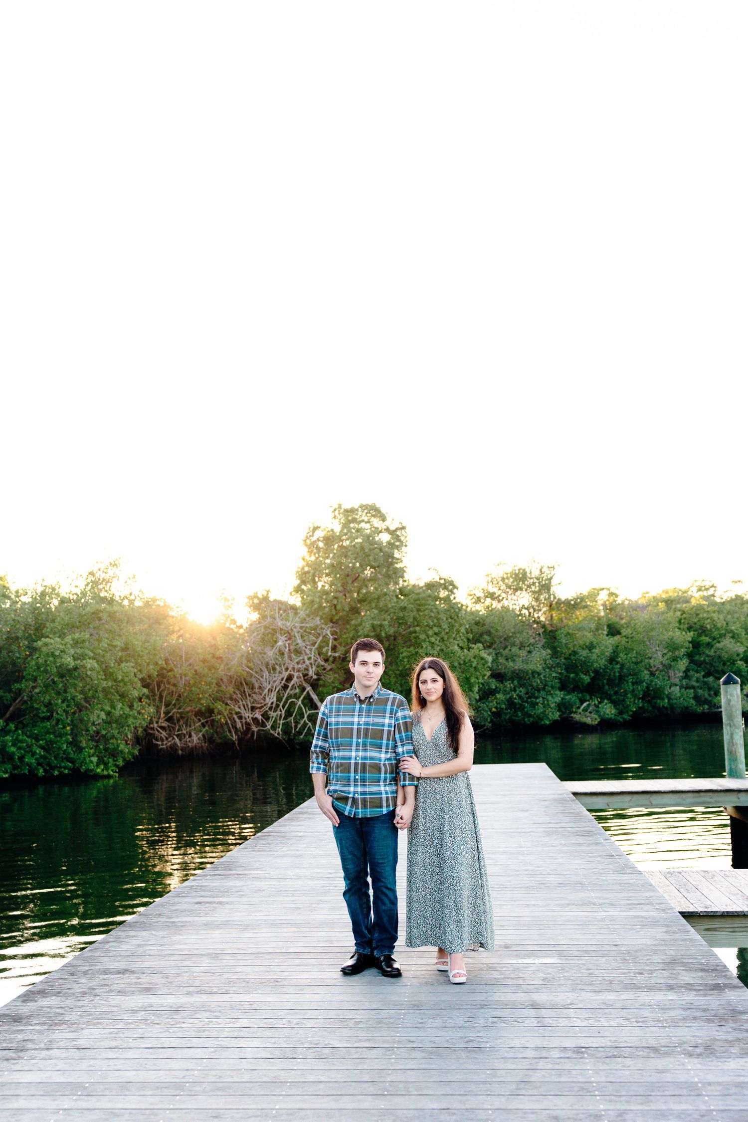 Couple standing on a wooden dock at sunset with lush green trees and calm water in the background.