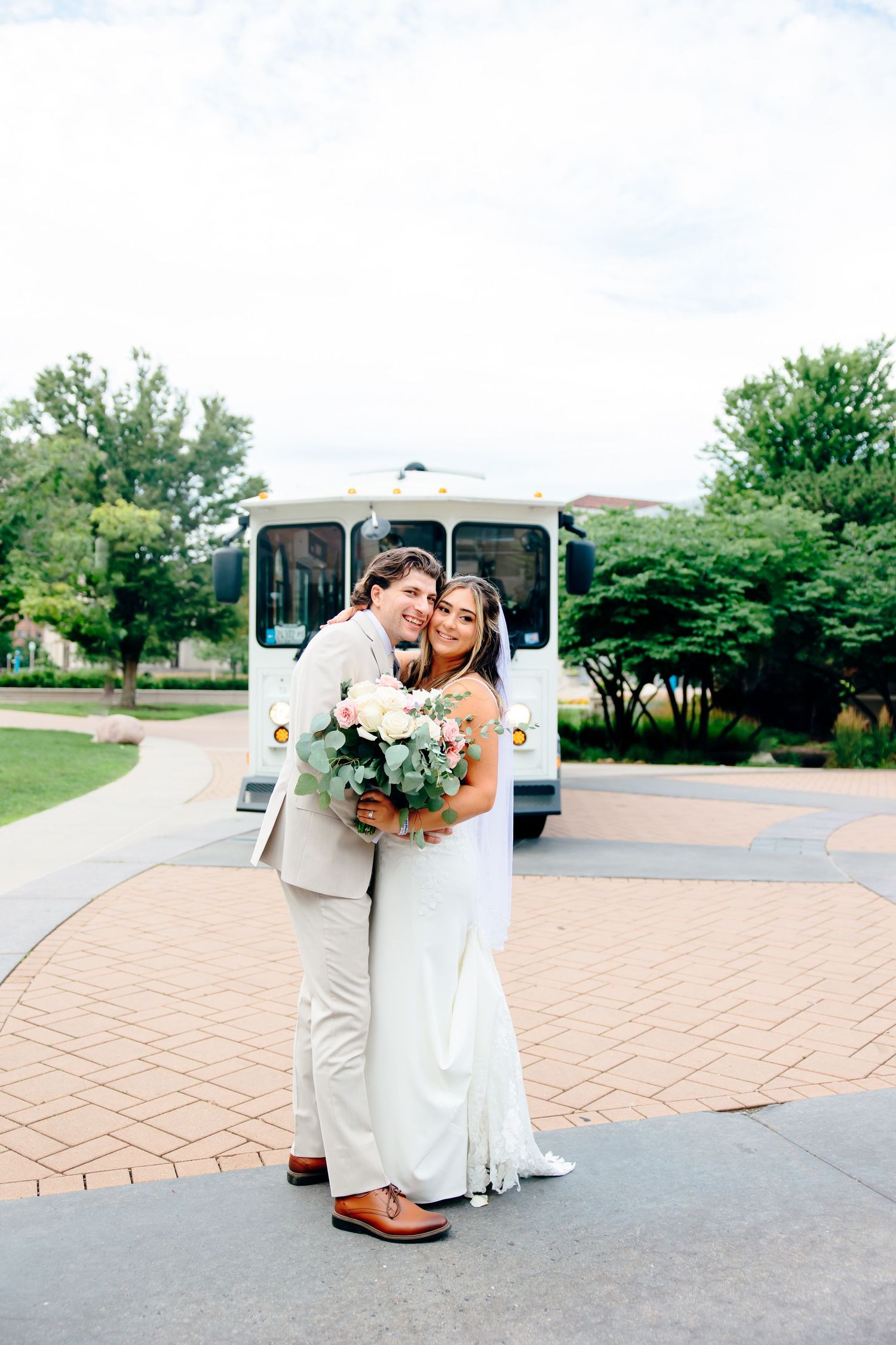 Bride and groom embracing and smiling in front of a white trolley bus on their wedding day.