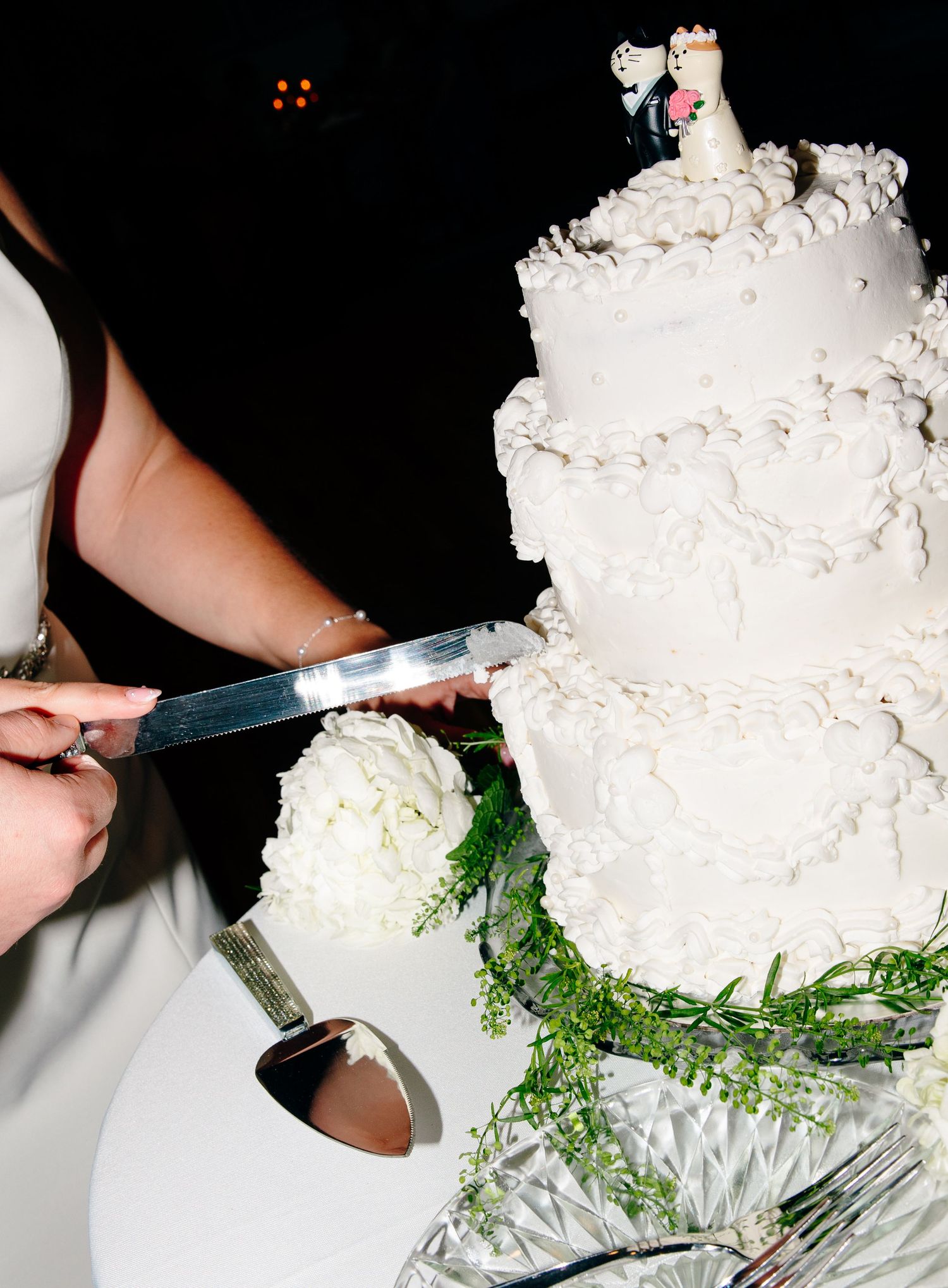 Bride cutting a three-tier white wedding cake topped with bride and groom figurines at reception.
