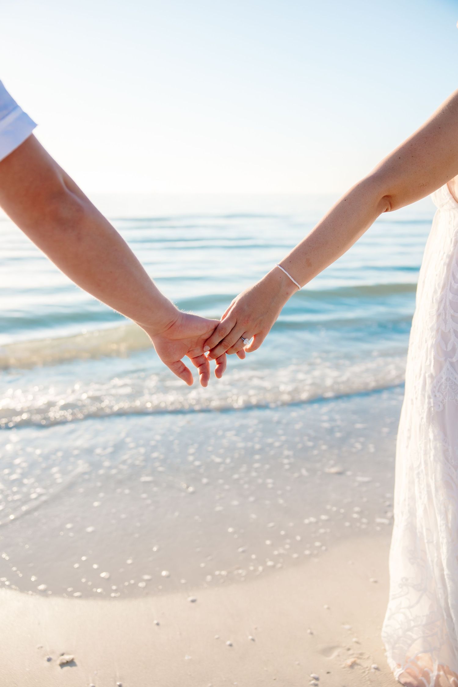 A couple holding hands while walking on a sandy beach with ocean waves in the background.
