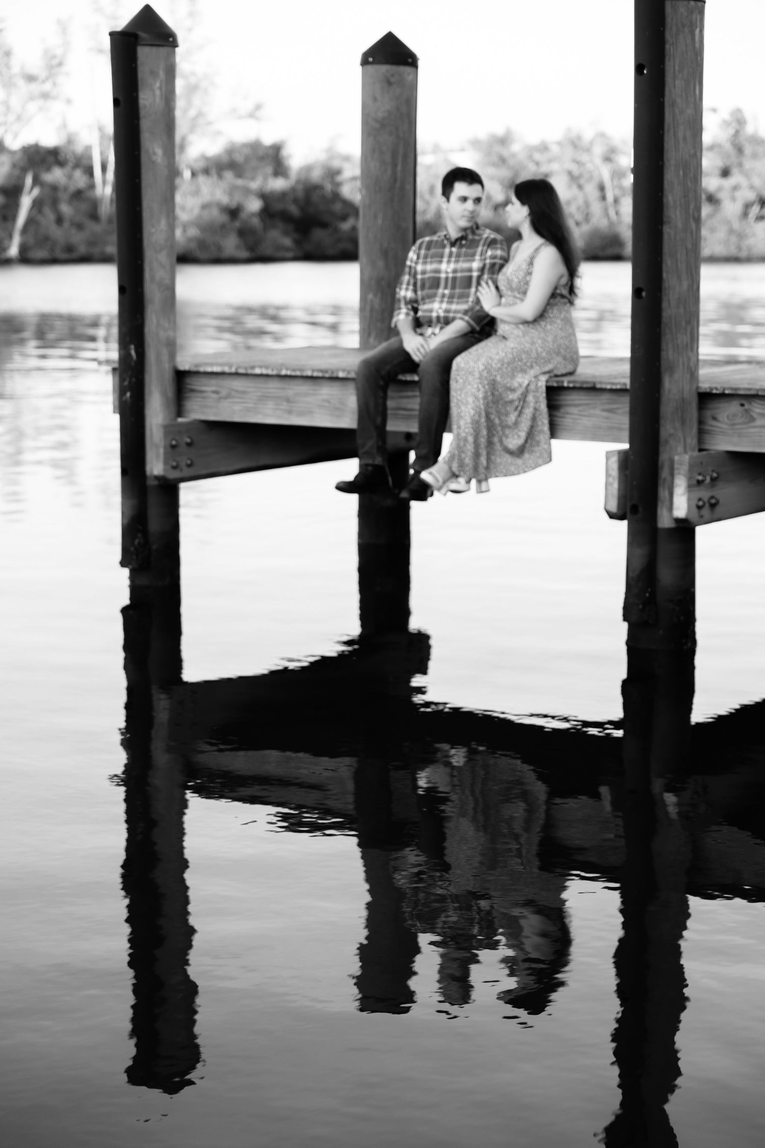 Couple sitting on a wooden dock over calm water, reflected below, in a romantic black and white photo.