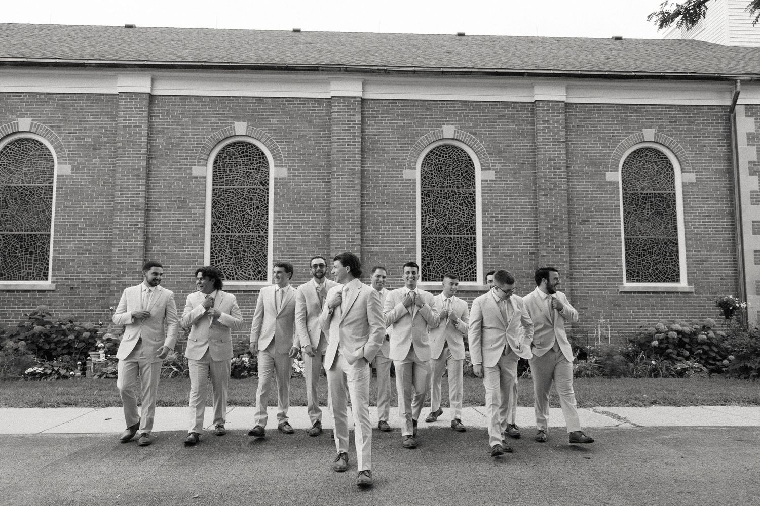 Groom and groomsmen in light suits walking together outside a brick church in a black and white wedding photo.