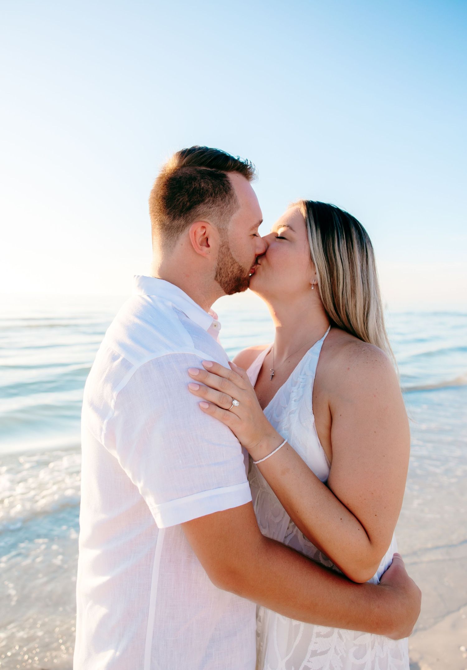 A romantic couple shares an intimate moment at the beach during sunset, dressed in white against the ocean backdrop.