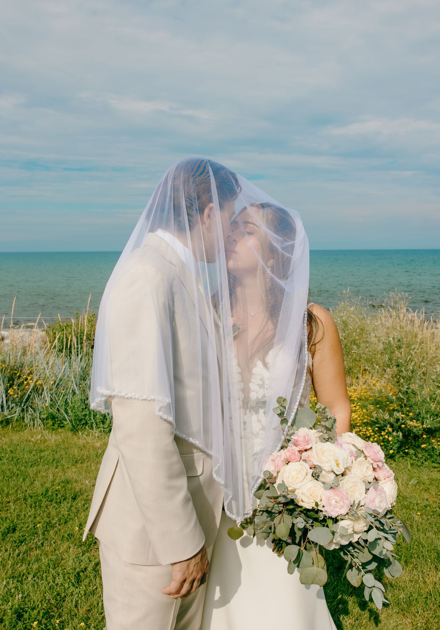 Romantic wedding photo by the ocean with couple under veil holding pink bouquet.