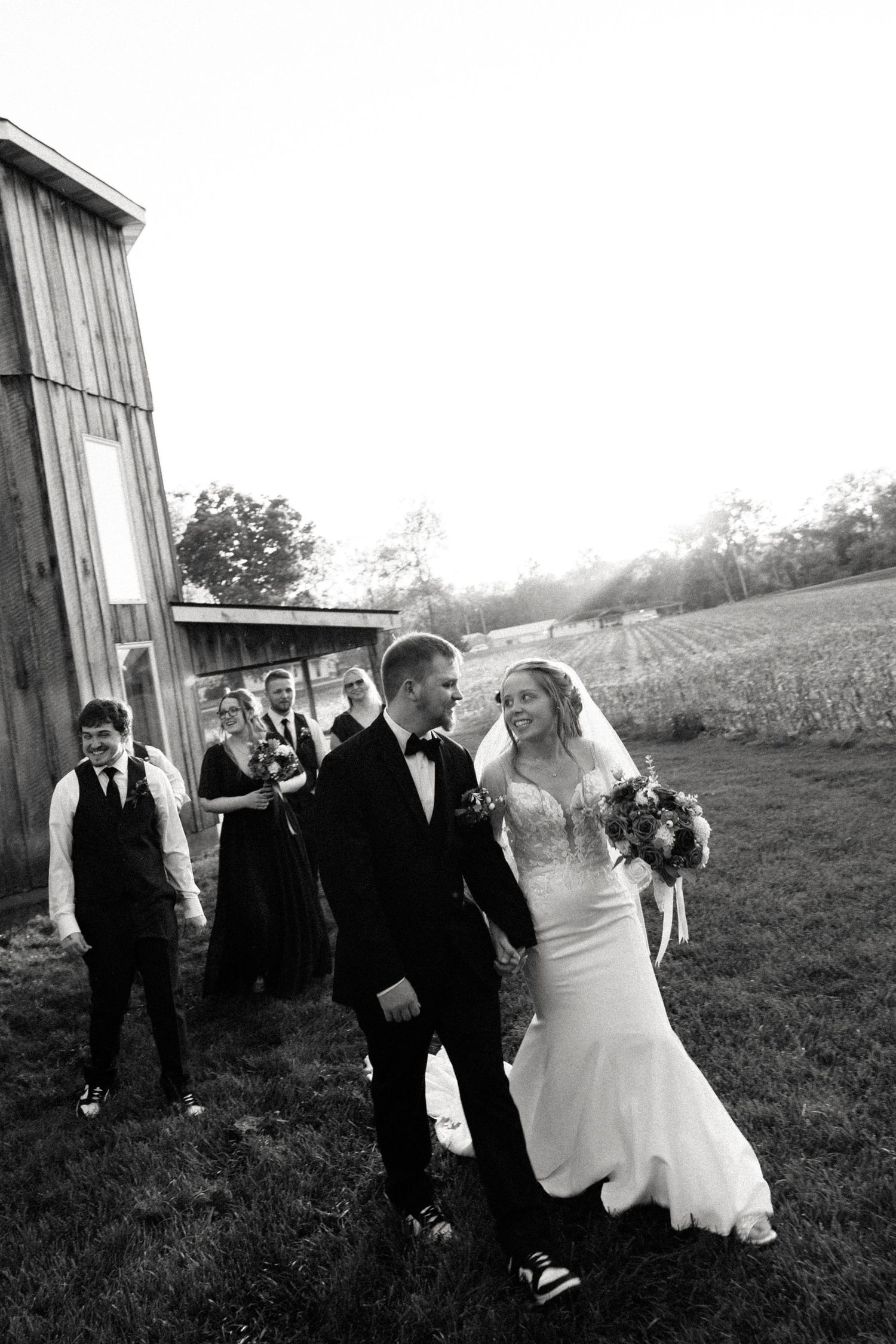 Black and white photo of a bride and groom walking together, smiling, with wedding party following near a rustic barn.