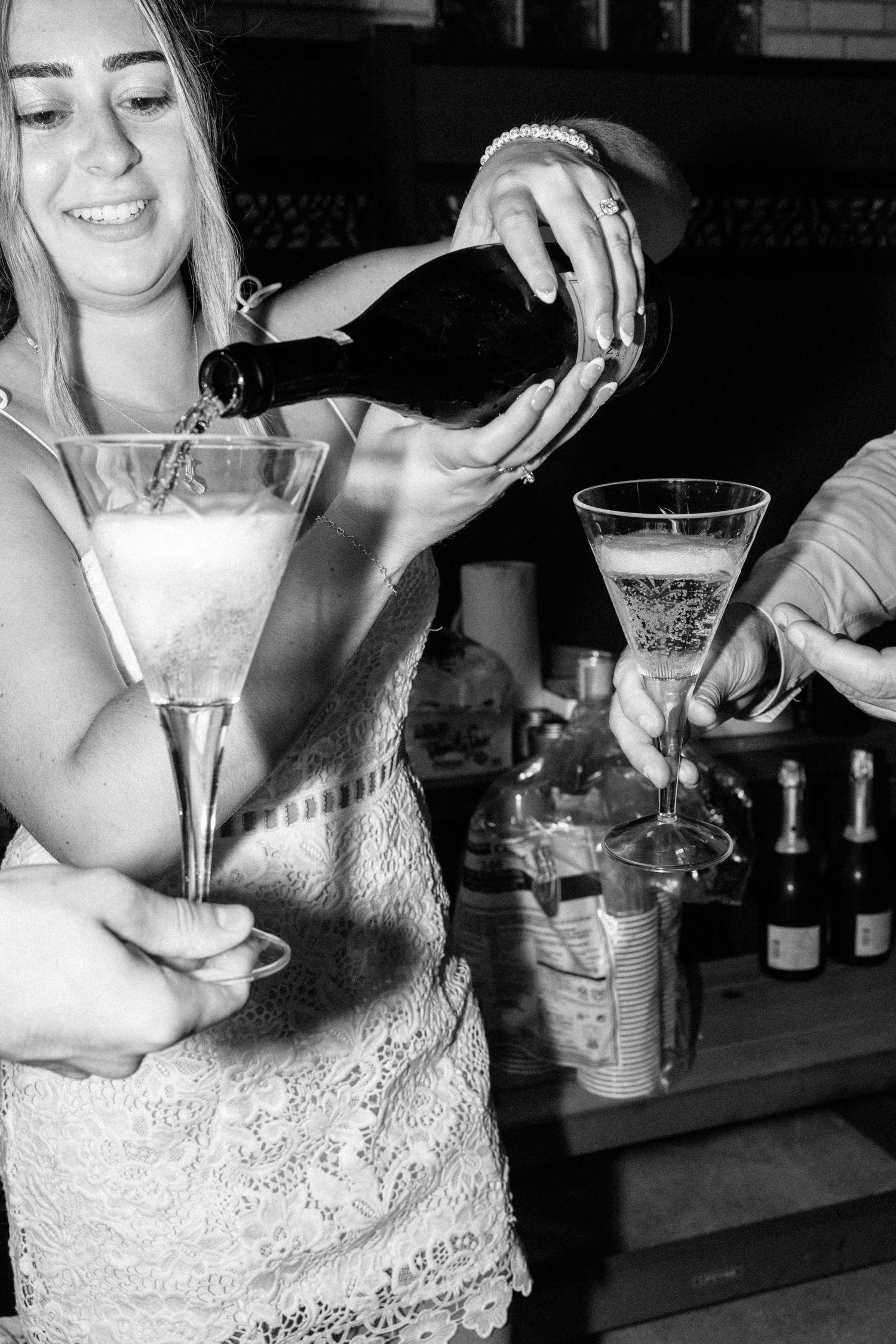 Black and white photo of a smiling woman pouring champagne into a large martini glass at a bar celebration.