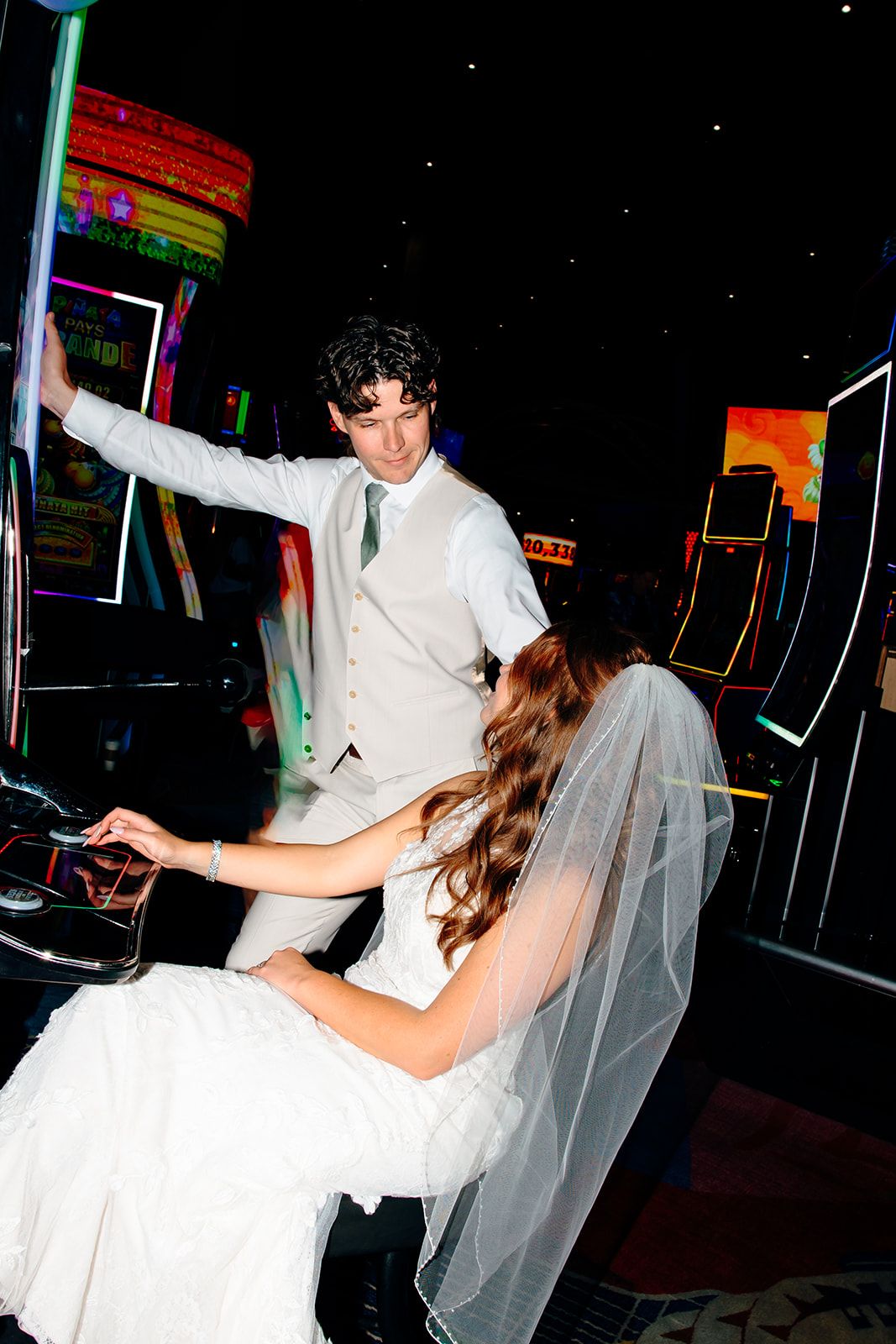 Bride in white gown and veil plays arcade games with groom in gray suit at a neon-lit wedding reception.