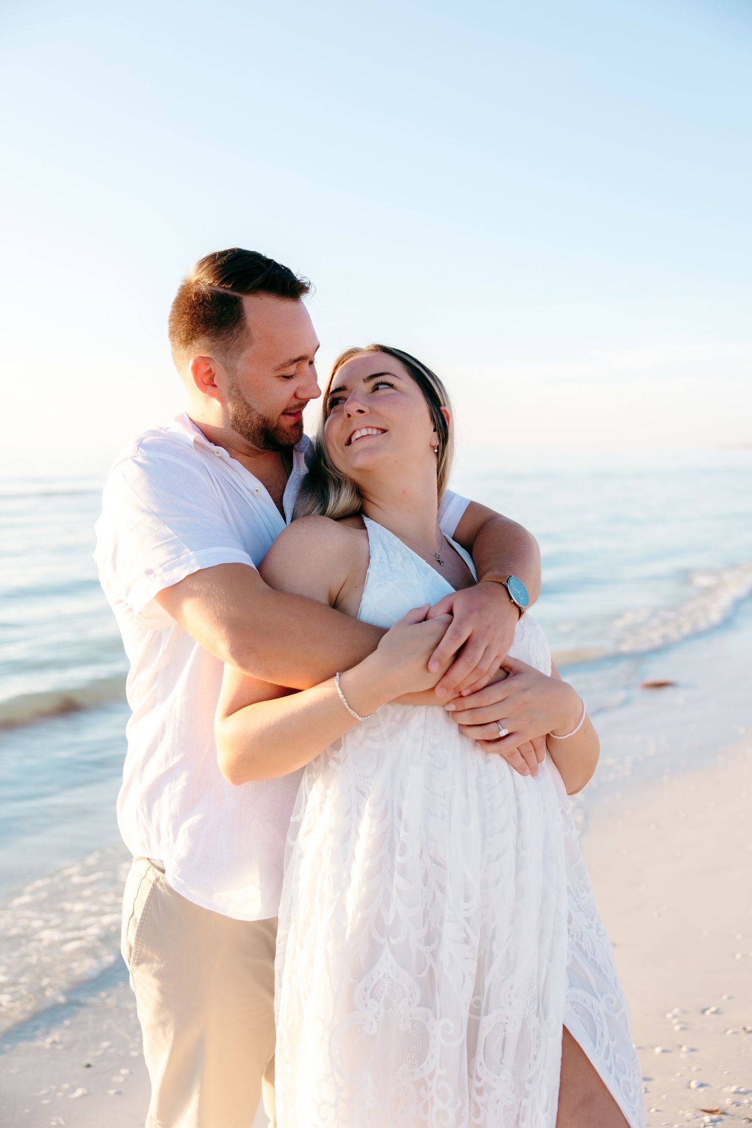 A romantic couple embraces on a sunny beach with waves in the background.