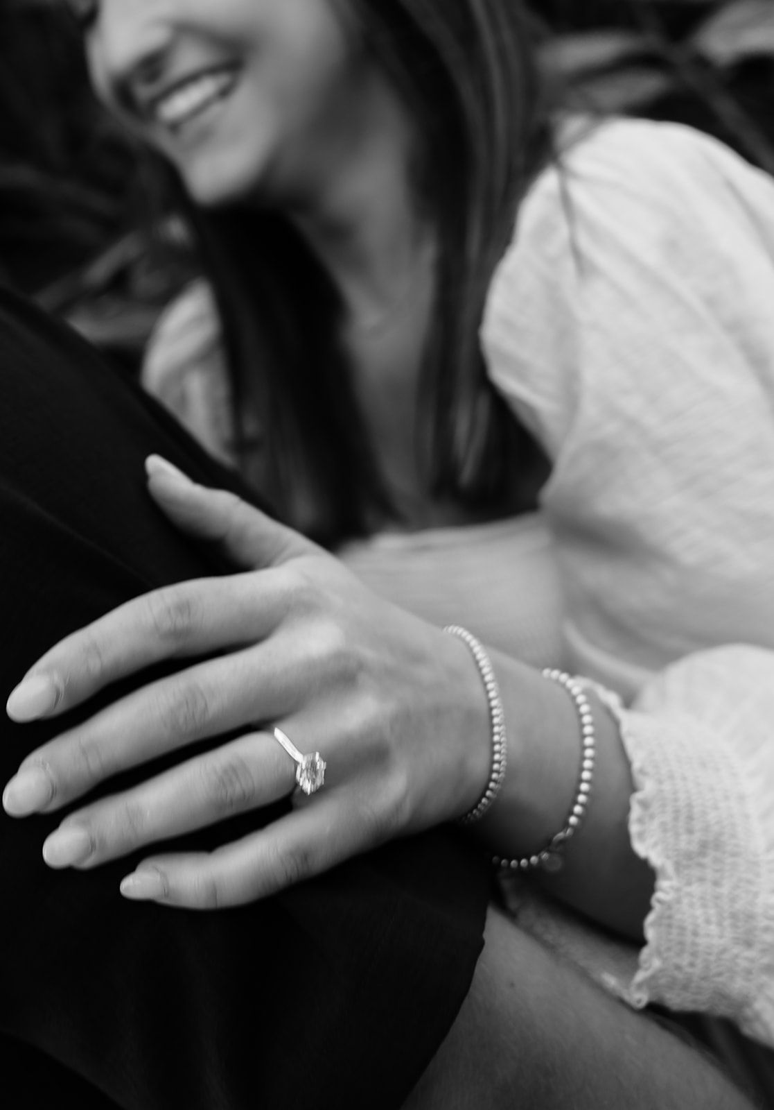 Black and white photo of a smiling woman displaying an engagement ring and beaded bracelet on her hand.