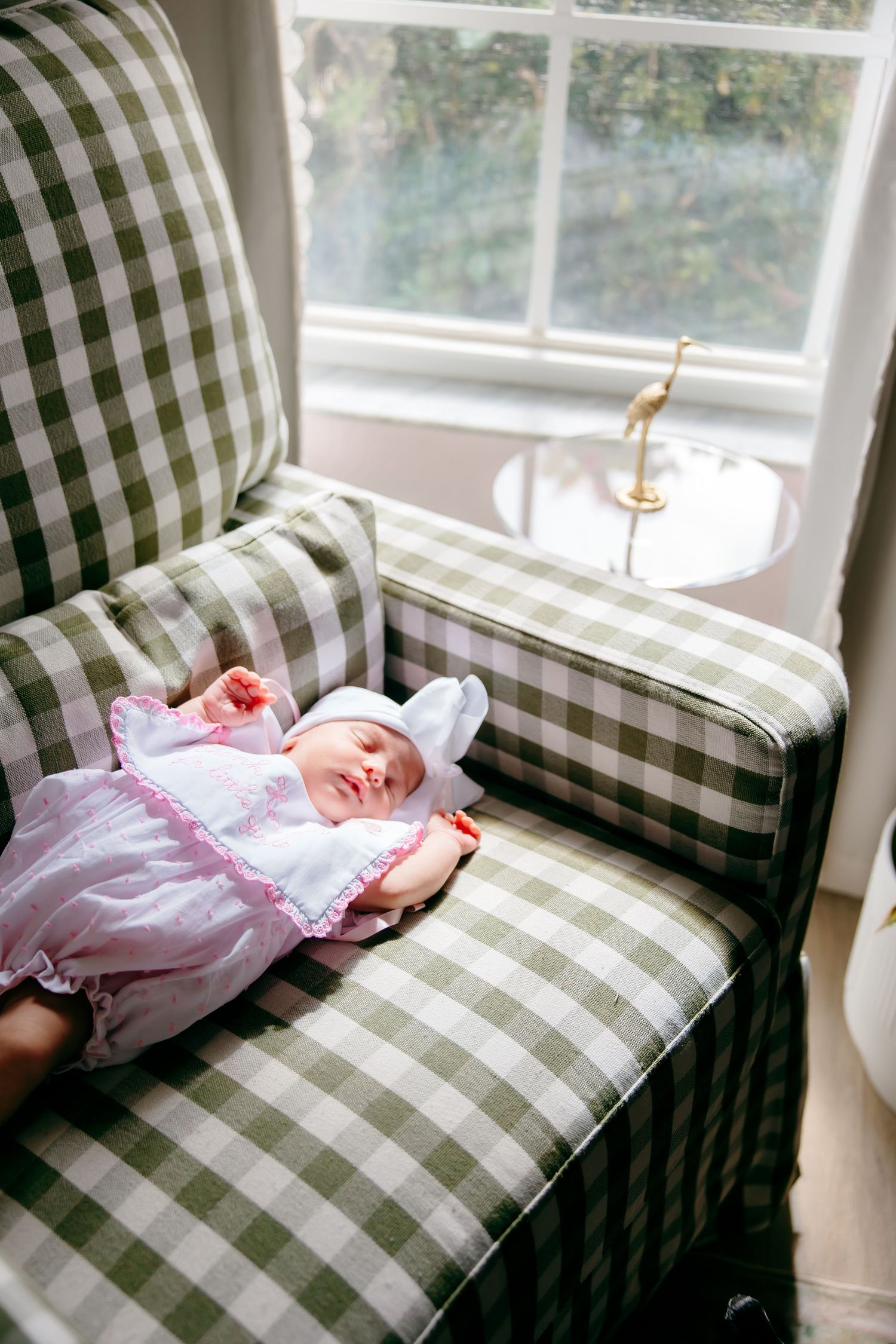 Newborn baby in pink dress sleeping peacefully on green gingham armchair near window.