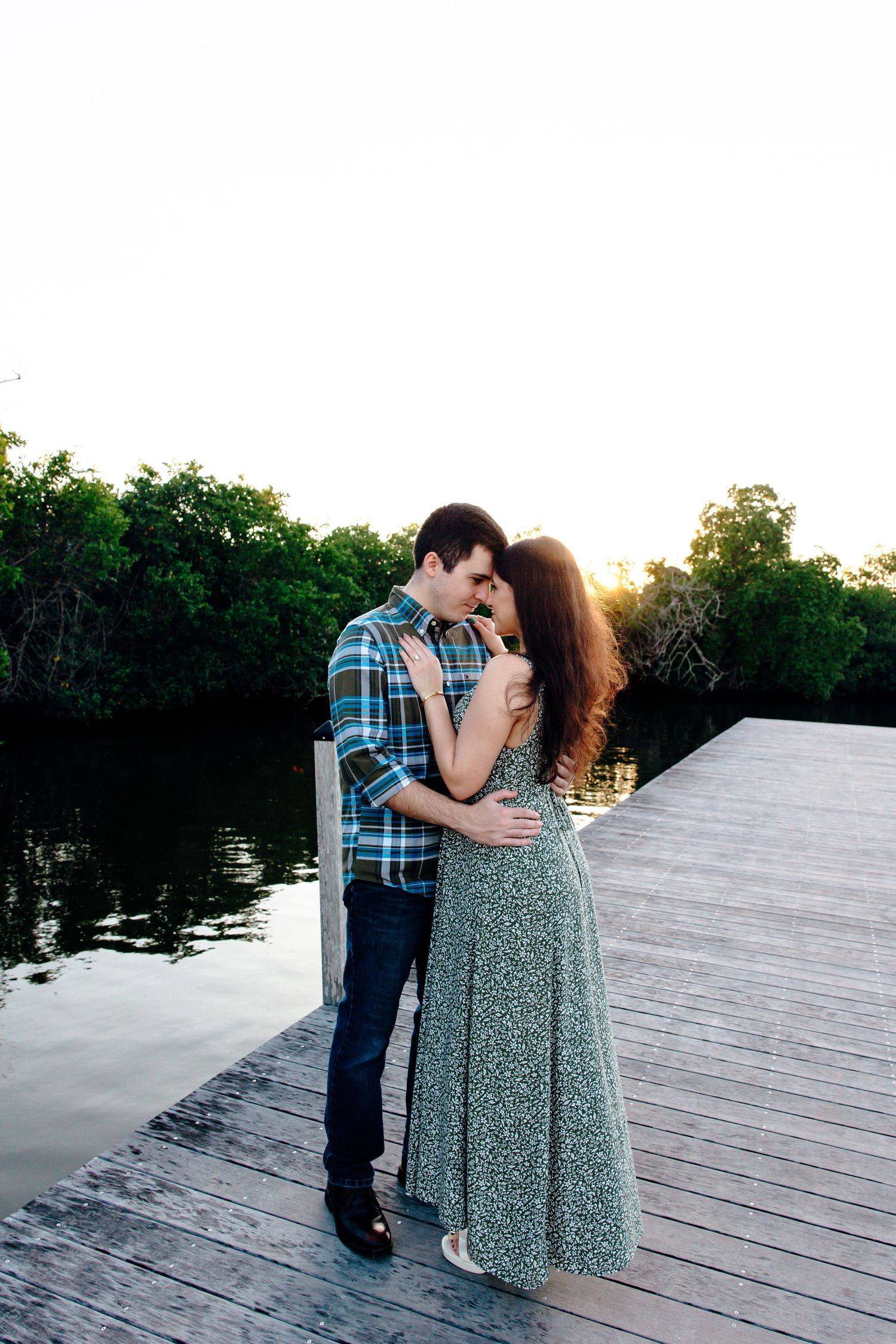 Couple embracing on a wooden dock at sunset, surrounded by lush green trees and calm water.