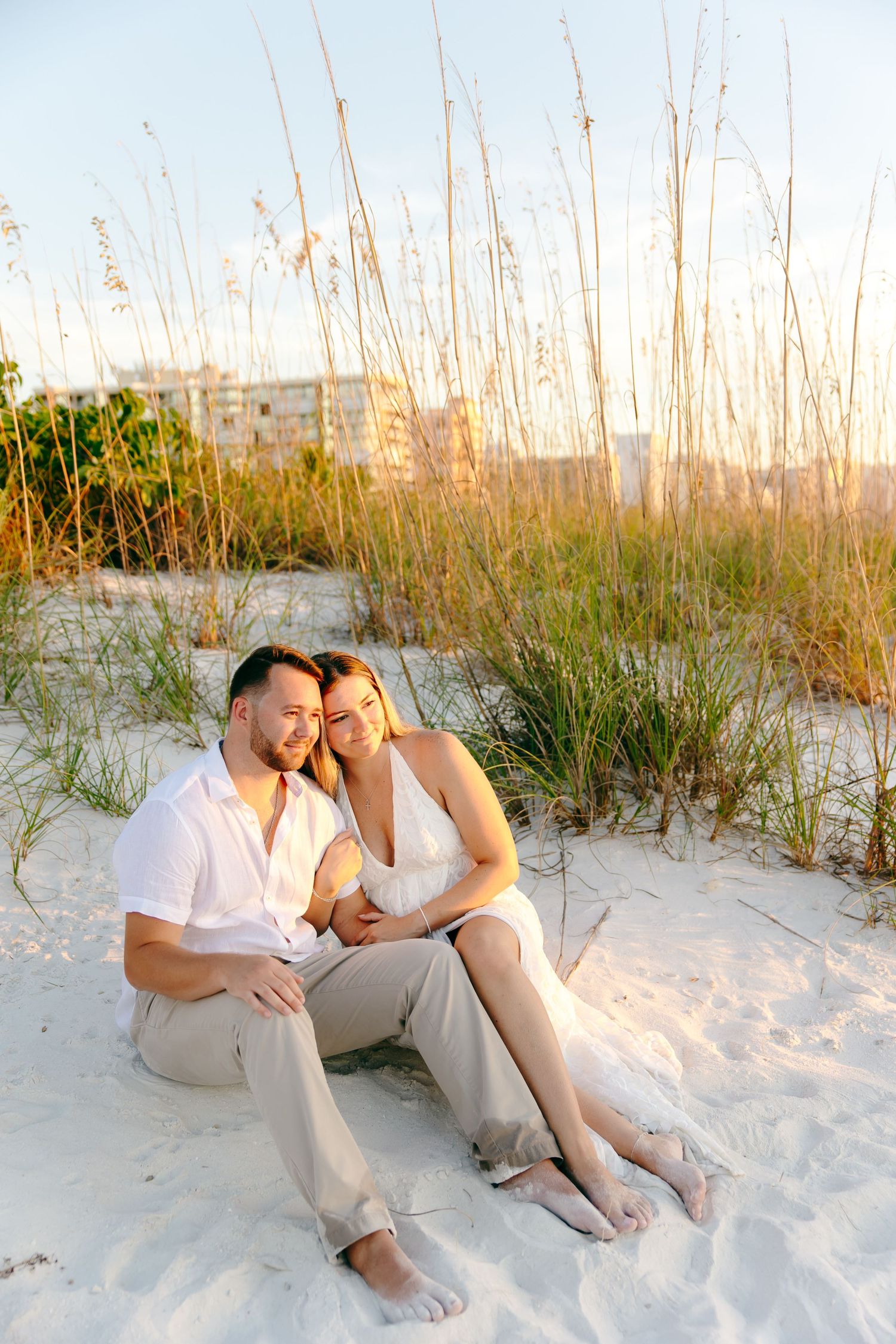 Couple sitting together on white sand beach dunes at sunset, surrounded by sea oats grass.