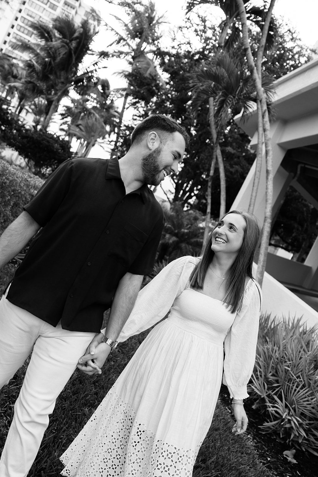 Black and white photo of a smiling couple holding hands and walking outdoors among palm trees.