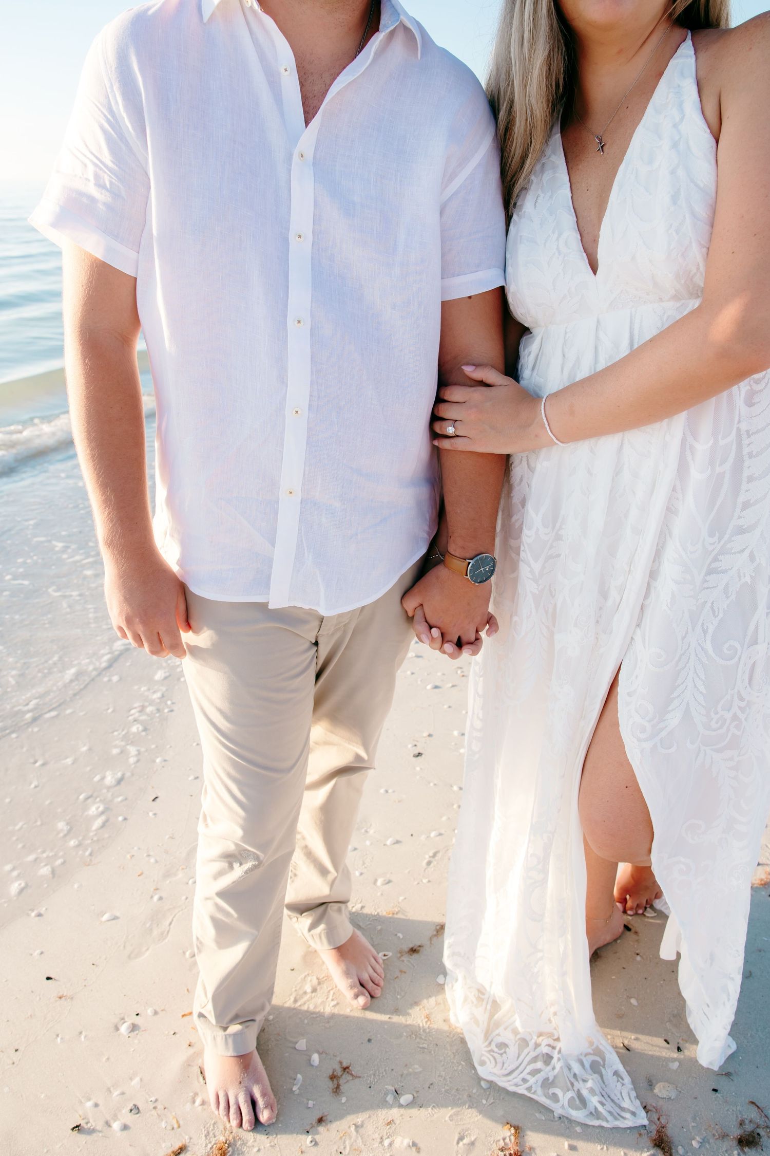 Couple in white outfits walking barefoot along a sandy beach shoreline at sunset.