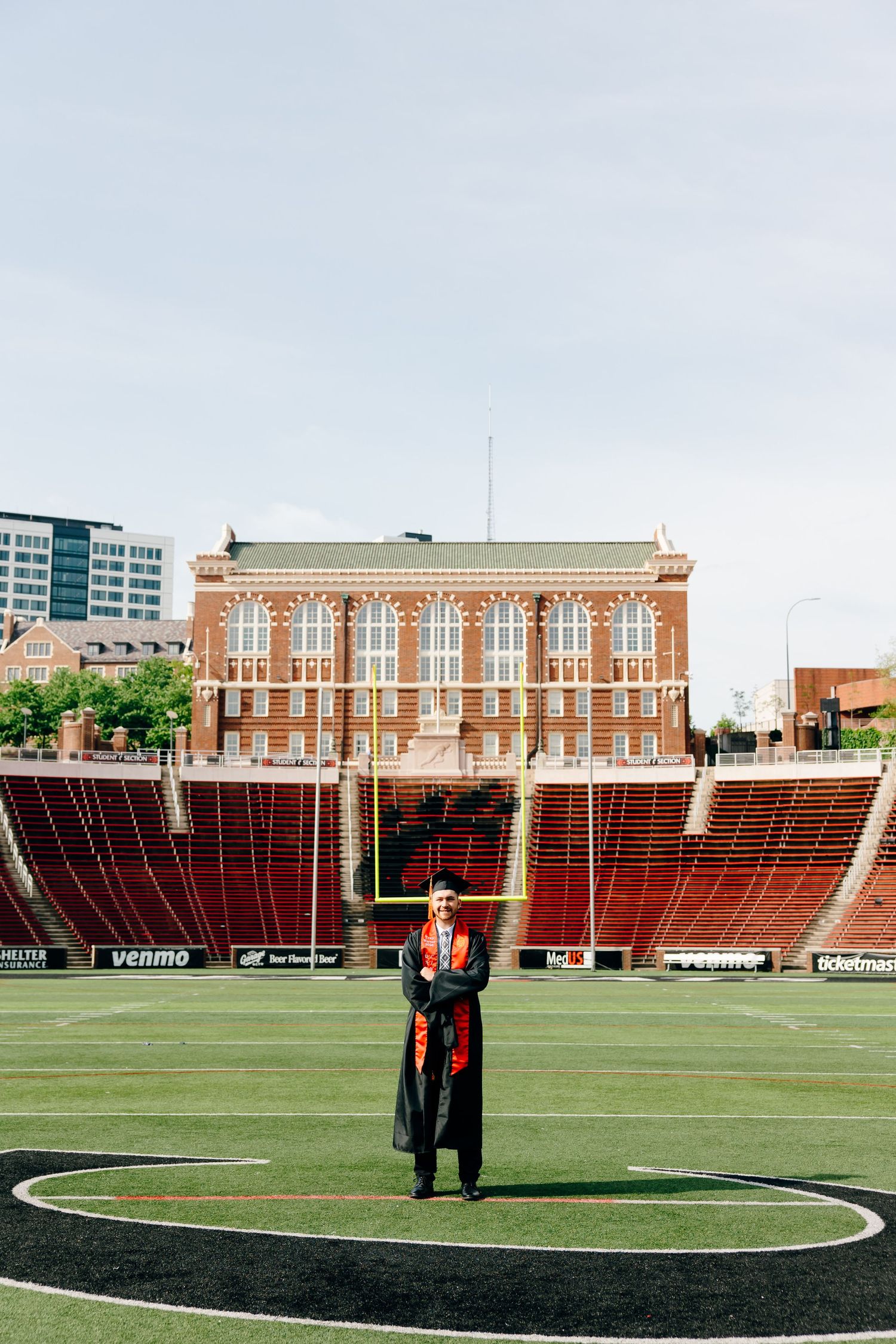 Graduate in cap and gown stands on football field with red stadium stands and brick building backdrop.