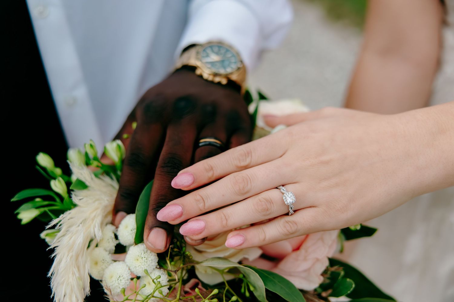 Interracial couple's hands showing wedding rings resting on bridal bouquet with white flowers.