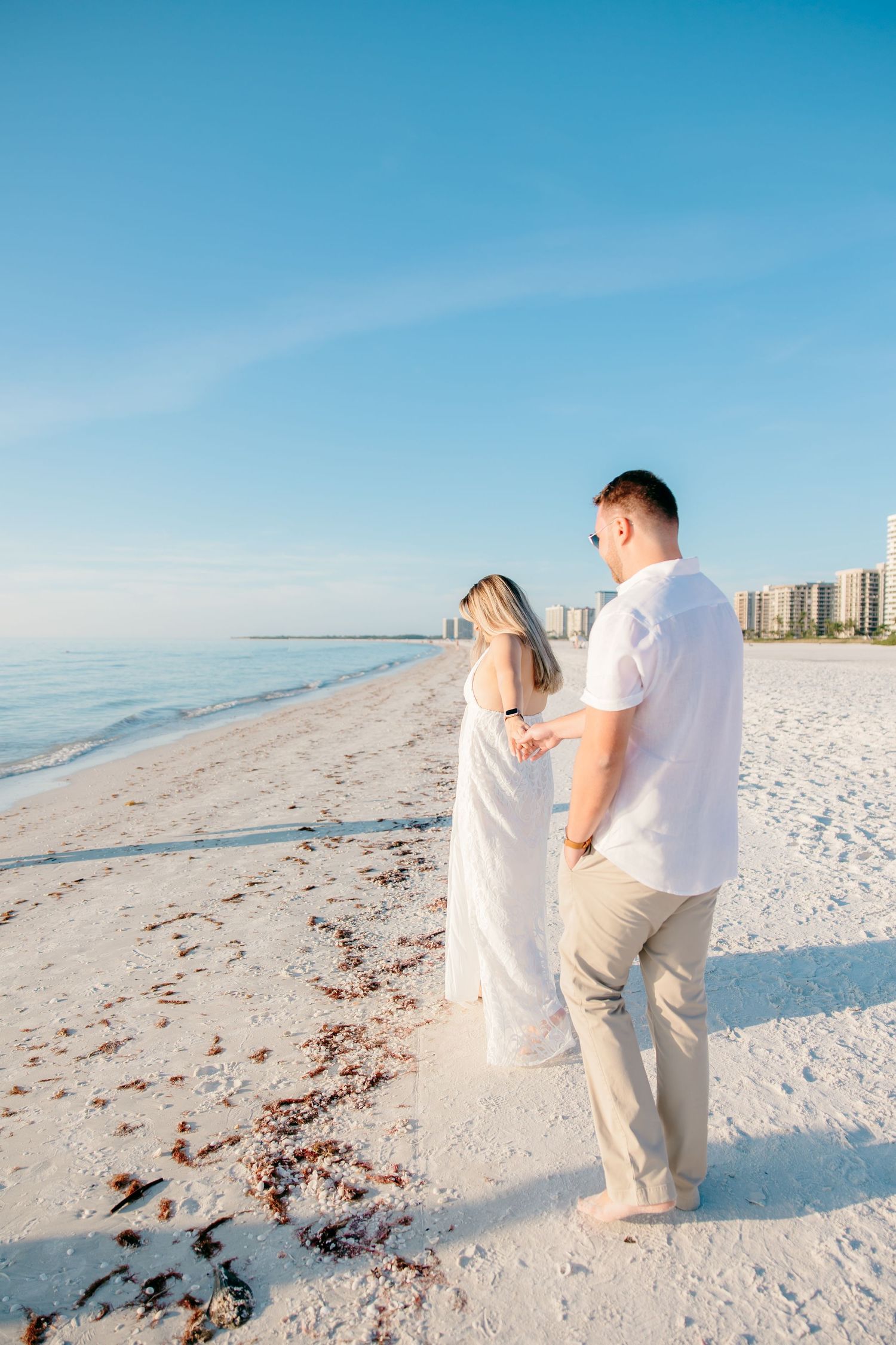 A couple walks hand in hand along a pristine white sandy beach with crystal blue waters and clear skies.