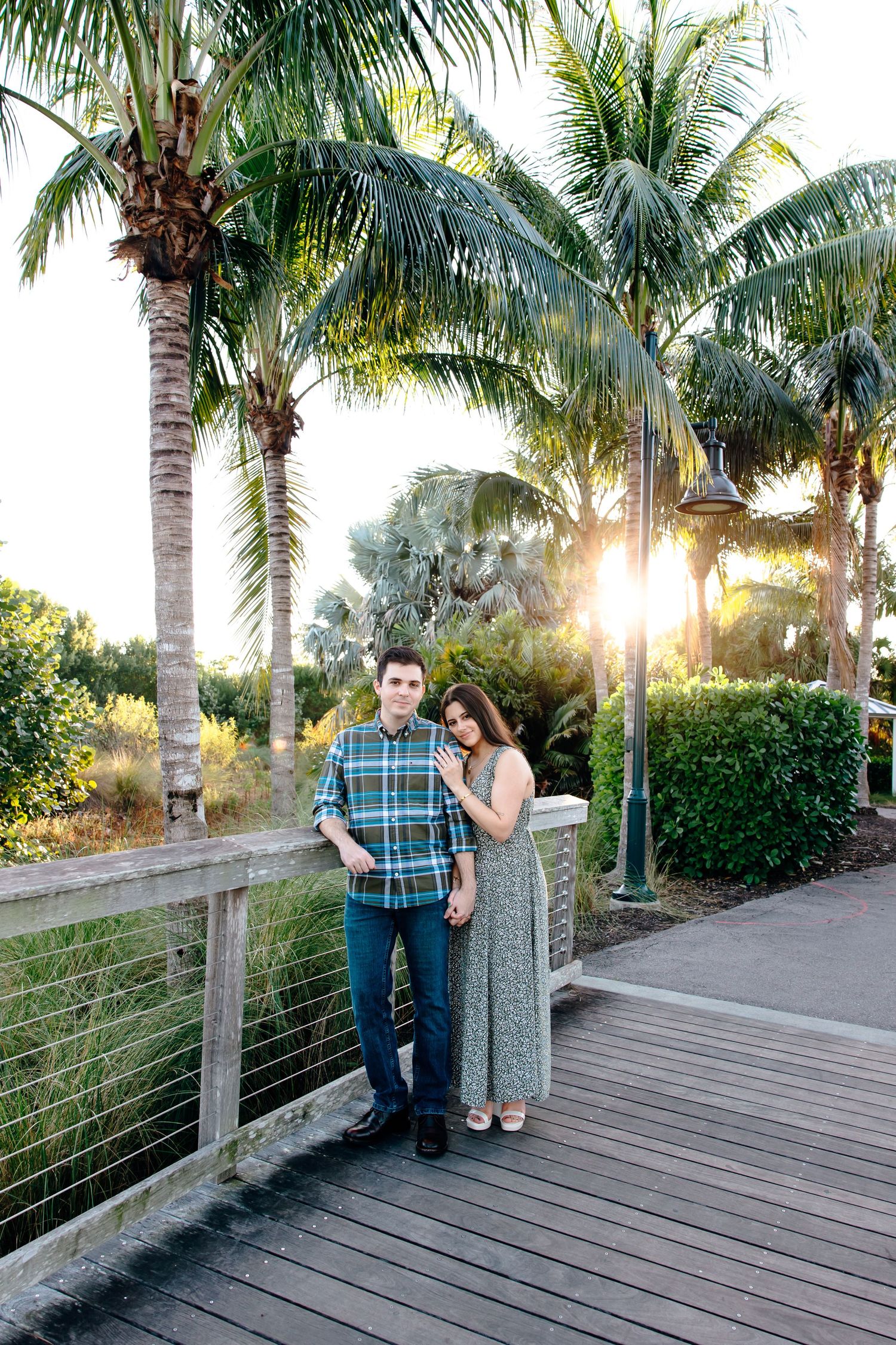 A couple poses on a wooden boardwalk surrounded by palm trees at golden hour in a tropical setting.