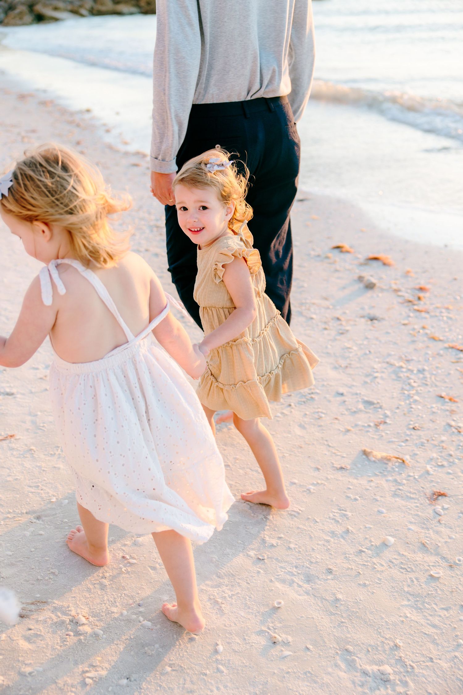 Two young girls in dresses walk barefoot on a sandy beach with an adult during golden hour.