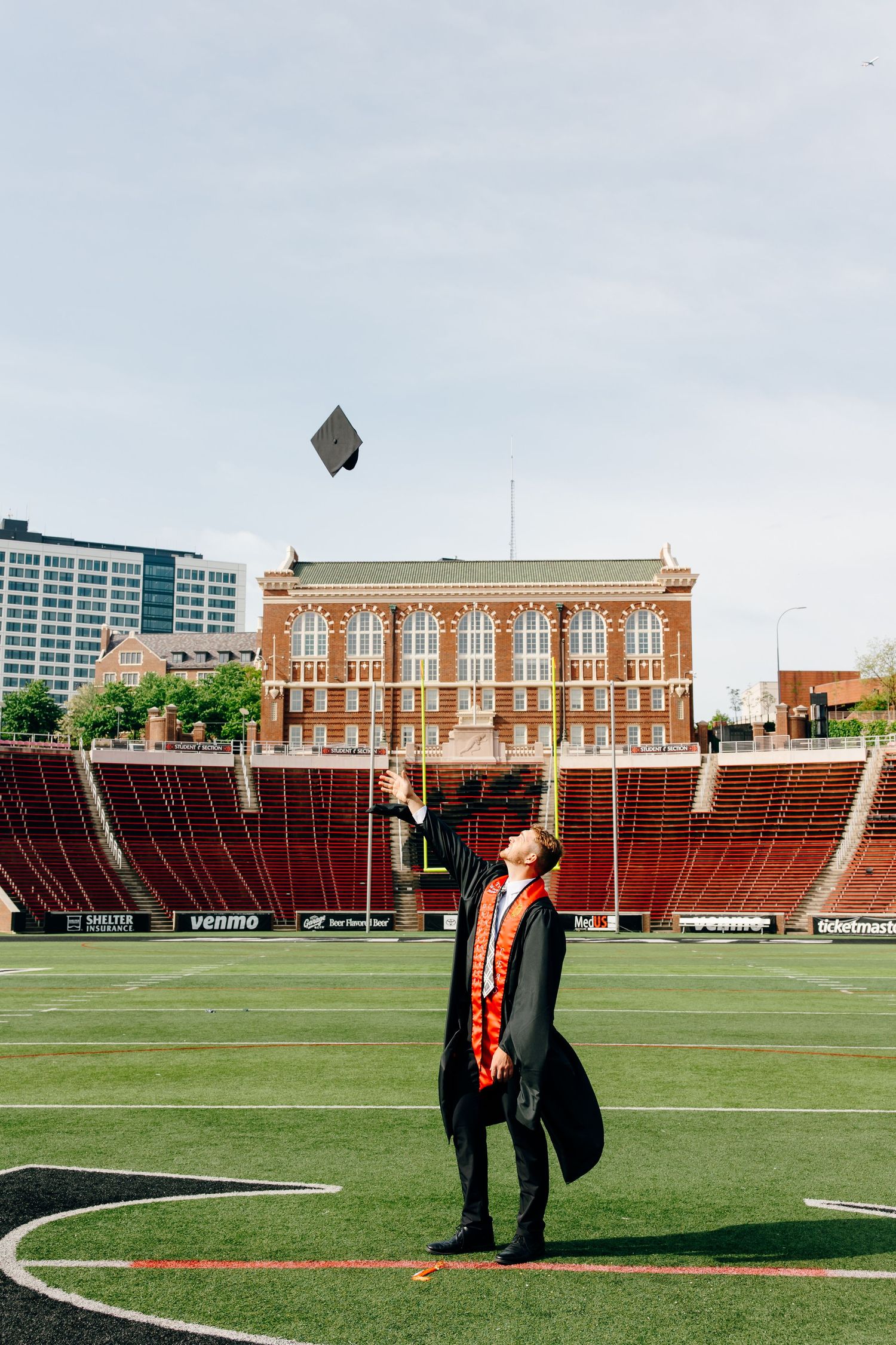 Graduate in cap and gown tosses mortarboard in air on football field with brick building backdrop.
