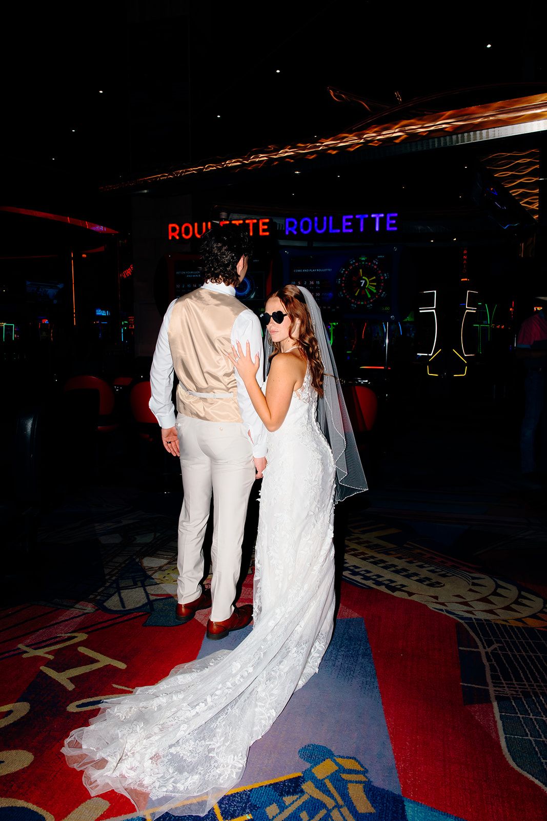 Bride and groom standing together in a casino near roulette signs, bride wearing lace gown with veil.