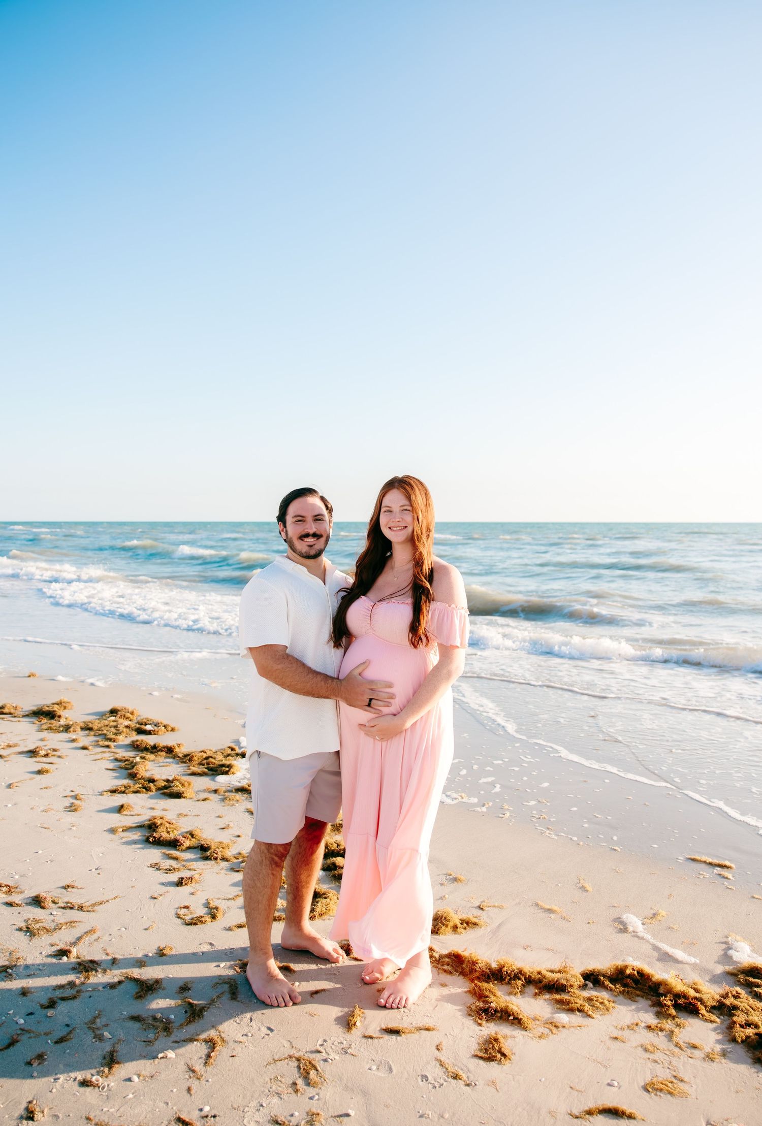 Couple posing on sunny beach, pregnant woman in pink dress with partner in white shirt and shorts.