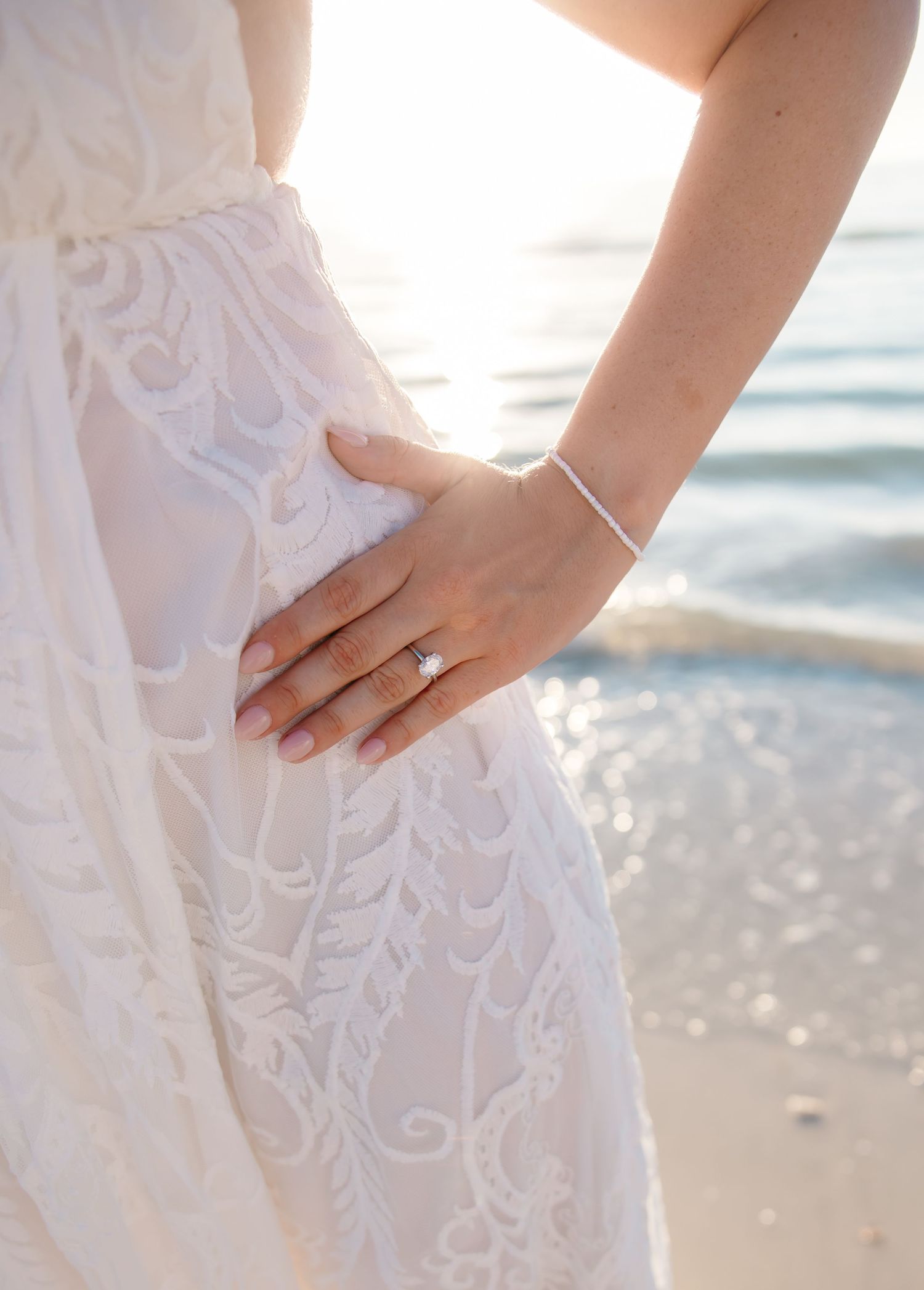 Close-up view of a diamond engagement ring being shown against a sunlit beach background with sparkling water.