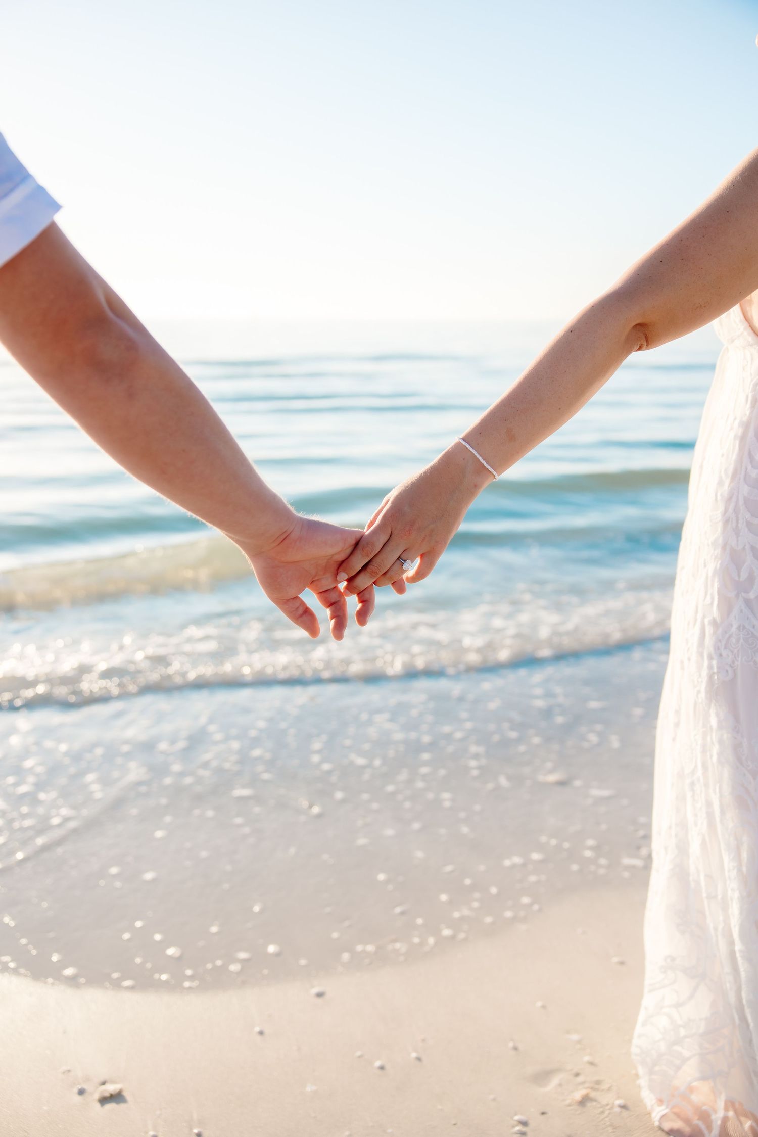 Couple holding hands on a sunny beach, woman wearing engagement ring and white dress near ocean waves.