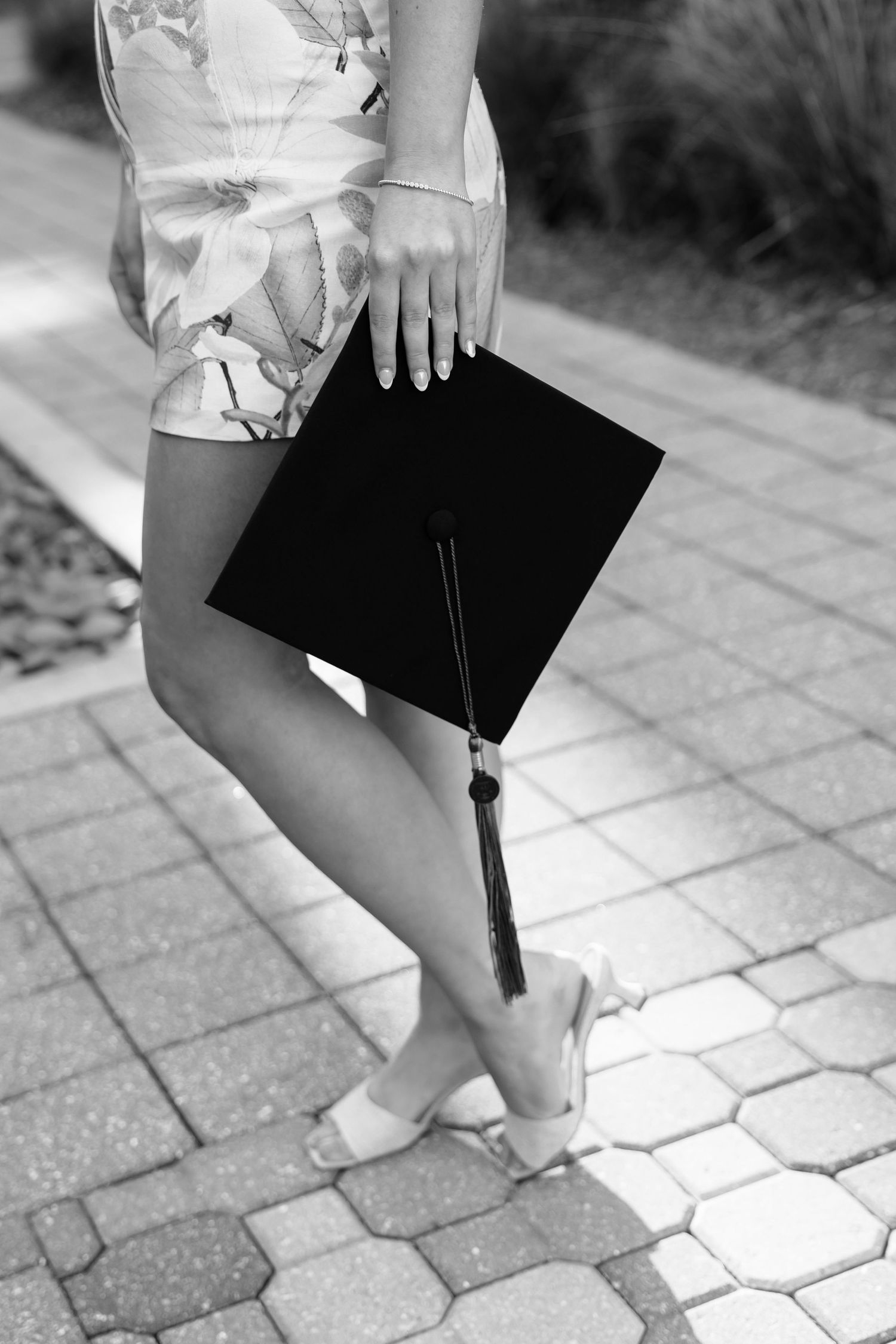 Black and white photo of a graduate holding a mortarboard cap while wearing a floral dress and heels on a brick path.