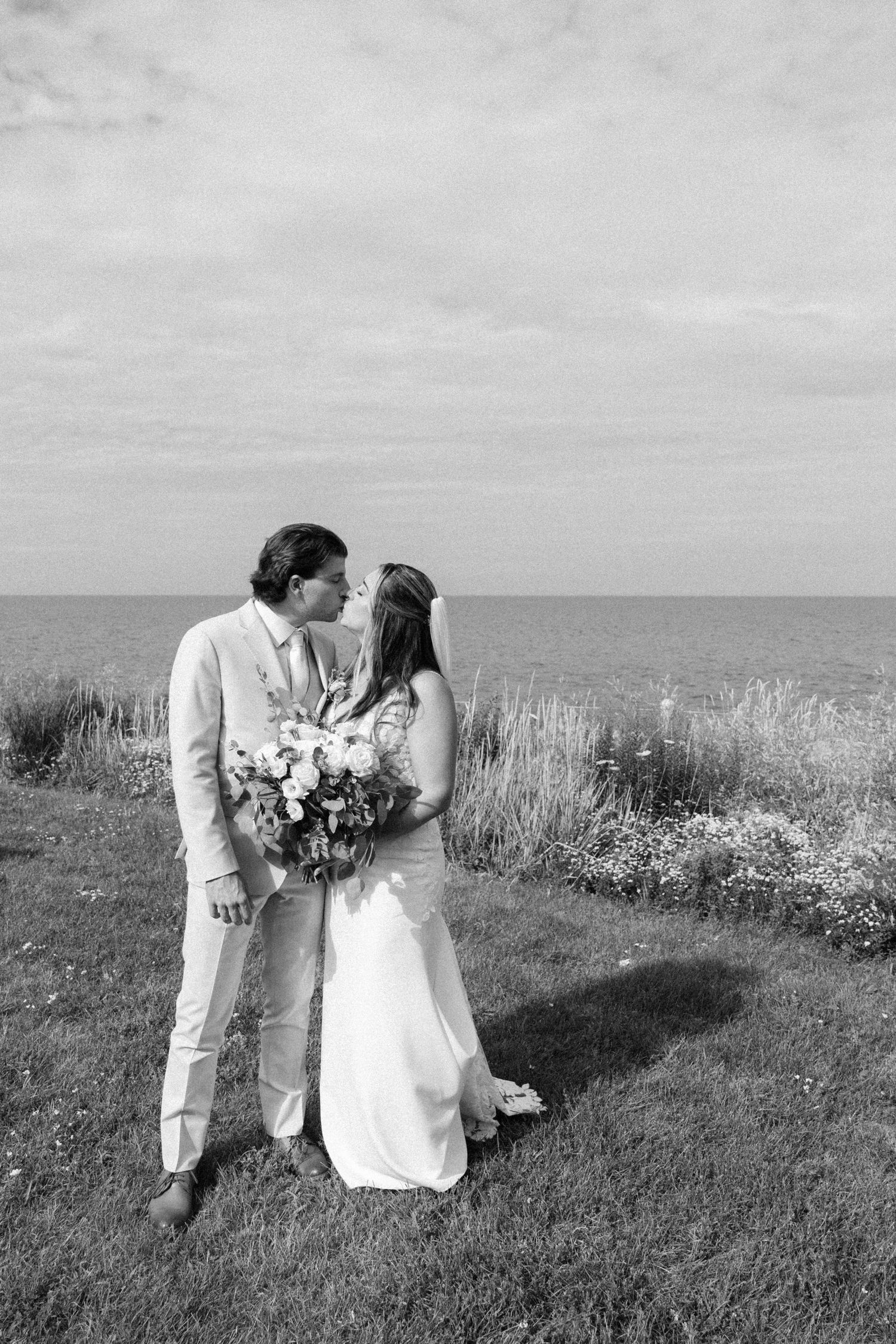 Black and white photo of a bride and groom kissing on a coastal cliff, bride holding a floral bouquet.