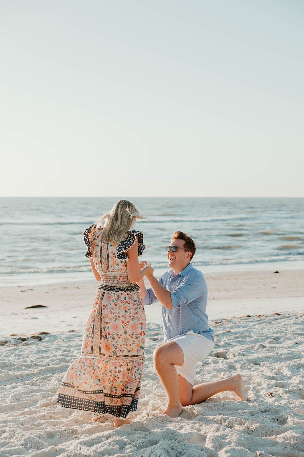 A romantic beach proposal scene with a couple on white sand as the sun sets over the ocean.