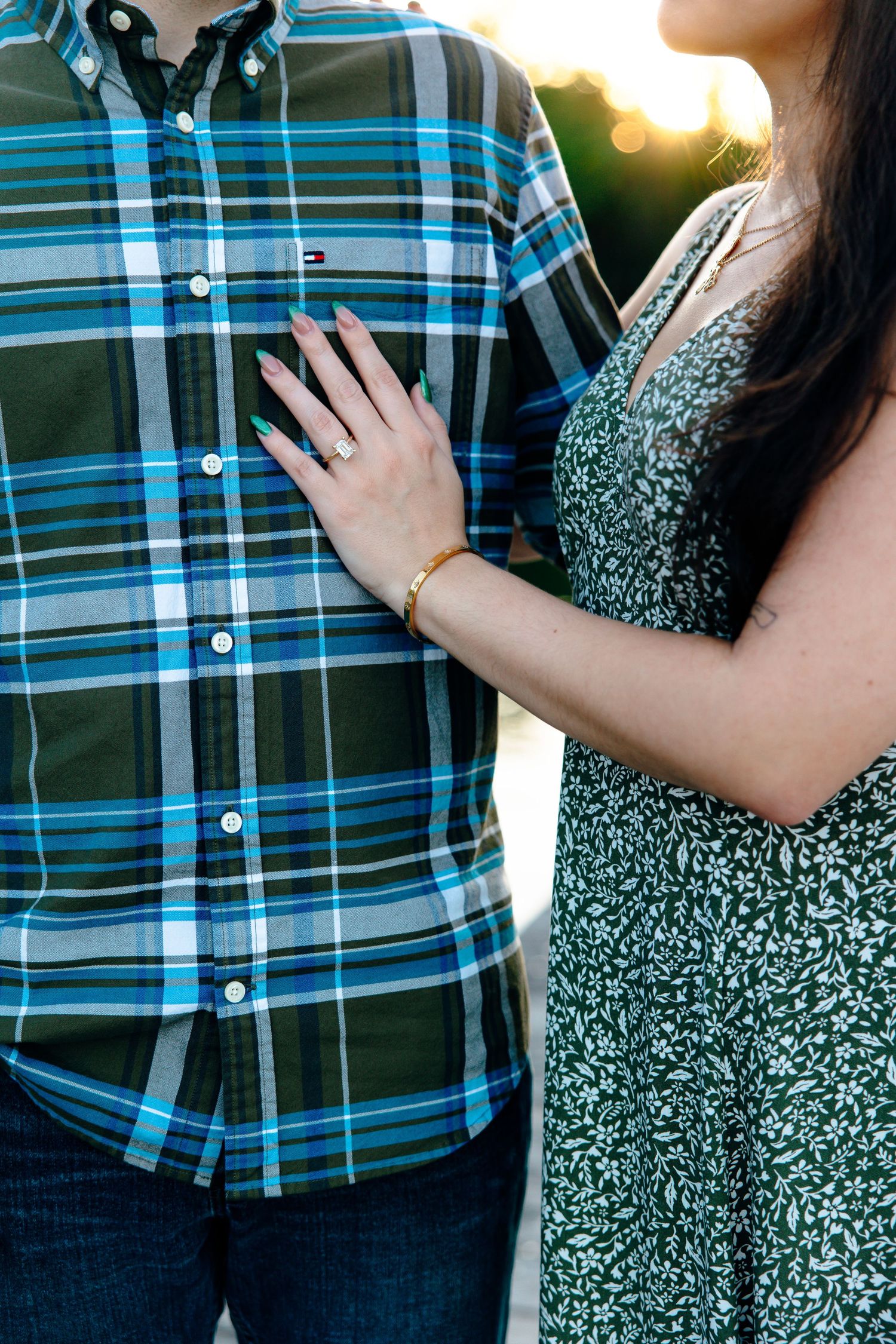 Couple standing close together, woman in floral dress with gold bracelet resting hand on man's plaid Tommy Hilfiger shirt.