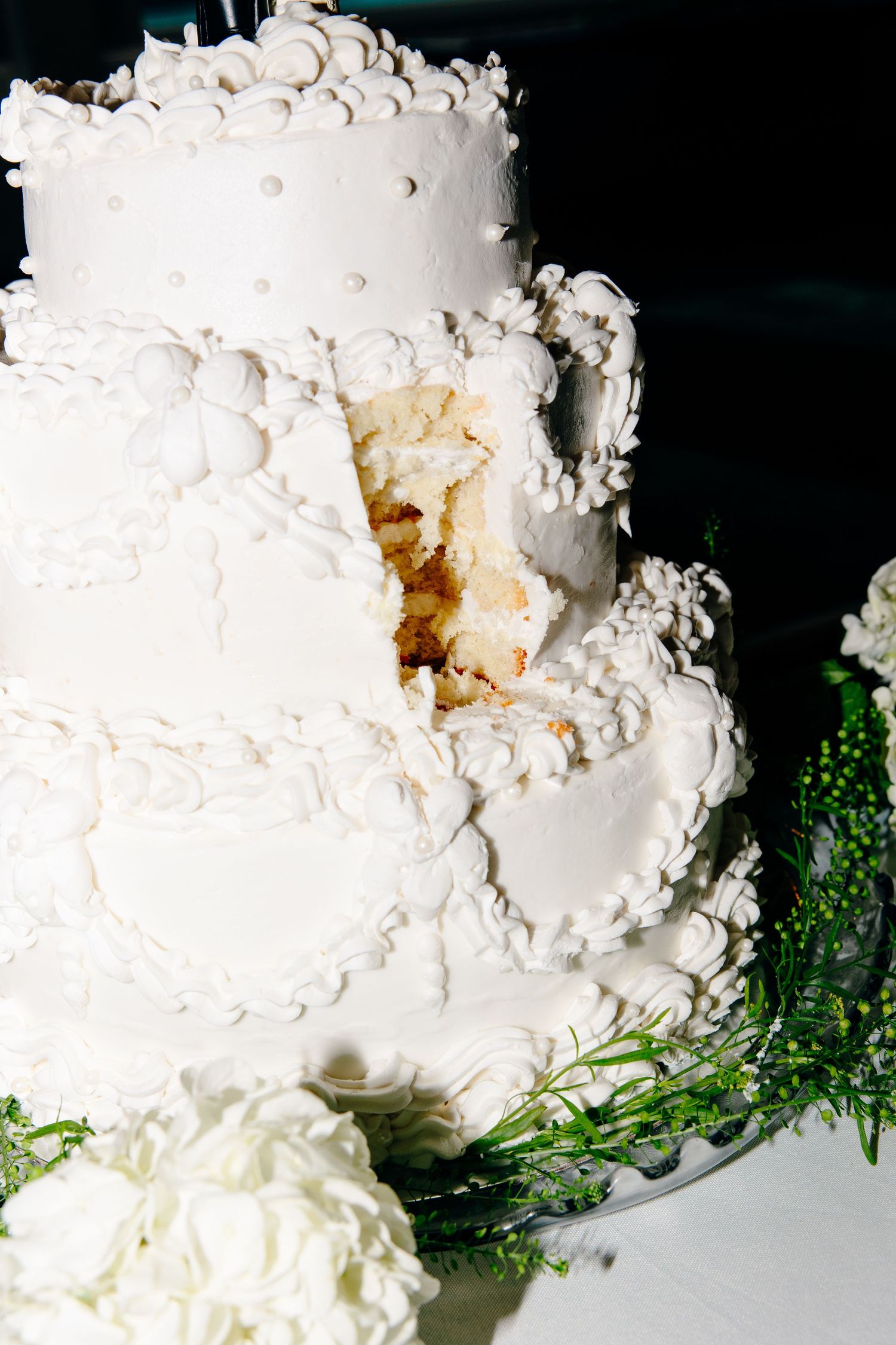 Elegant three-tier white wedding cake with intricate floral frosting details, partially cut to reveal moist interior layers.